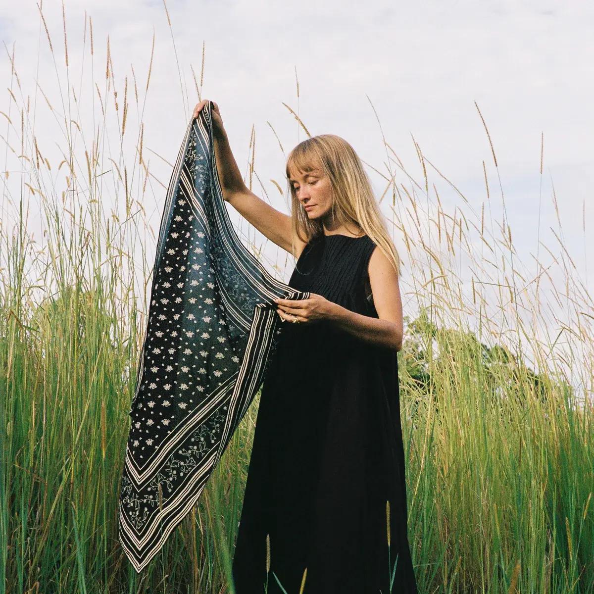 A blonde woman in a black dress holds up and examines a patterned scarf in a field of tall grass.