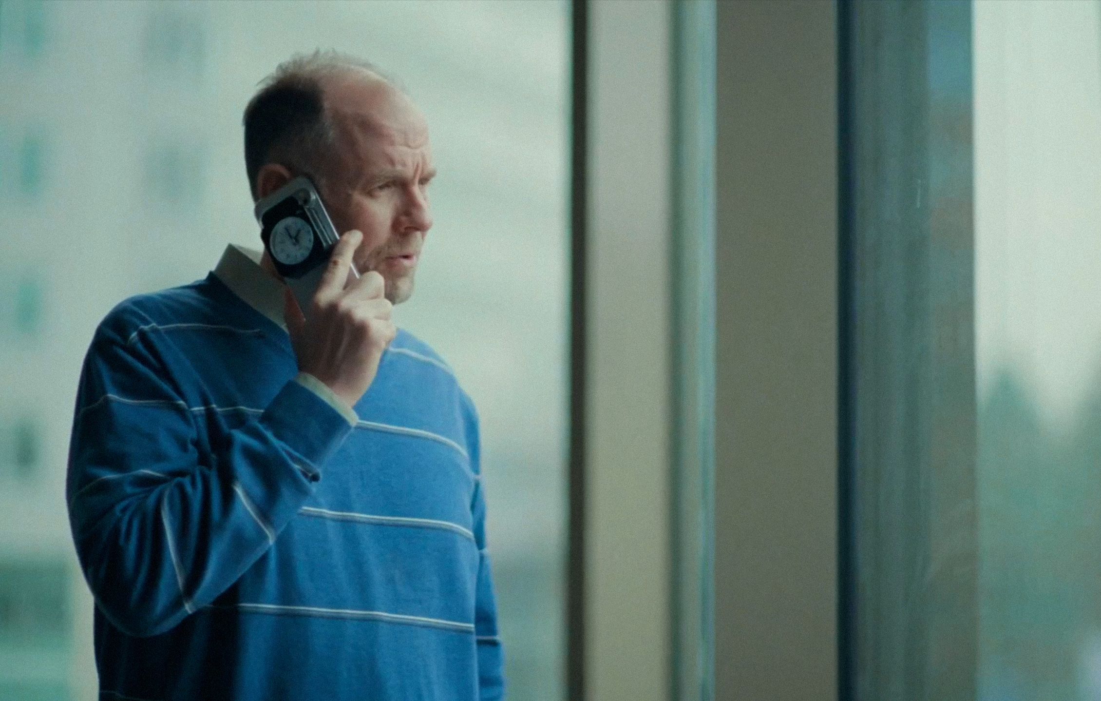 A man in a blue striped sweater talks on a cell phone with a small clock on its back, looking out a window.