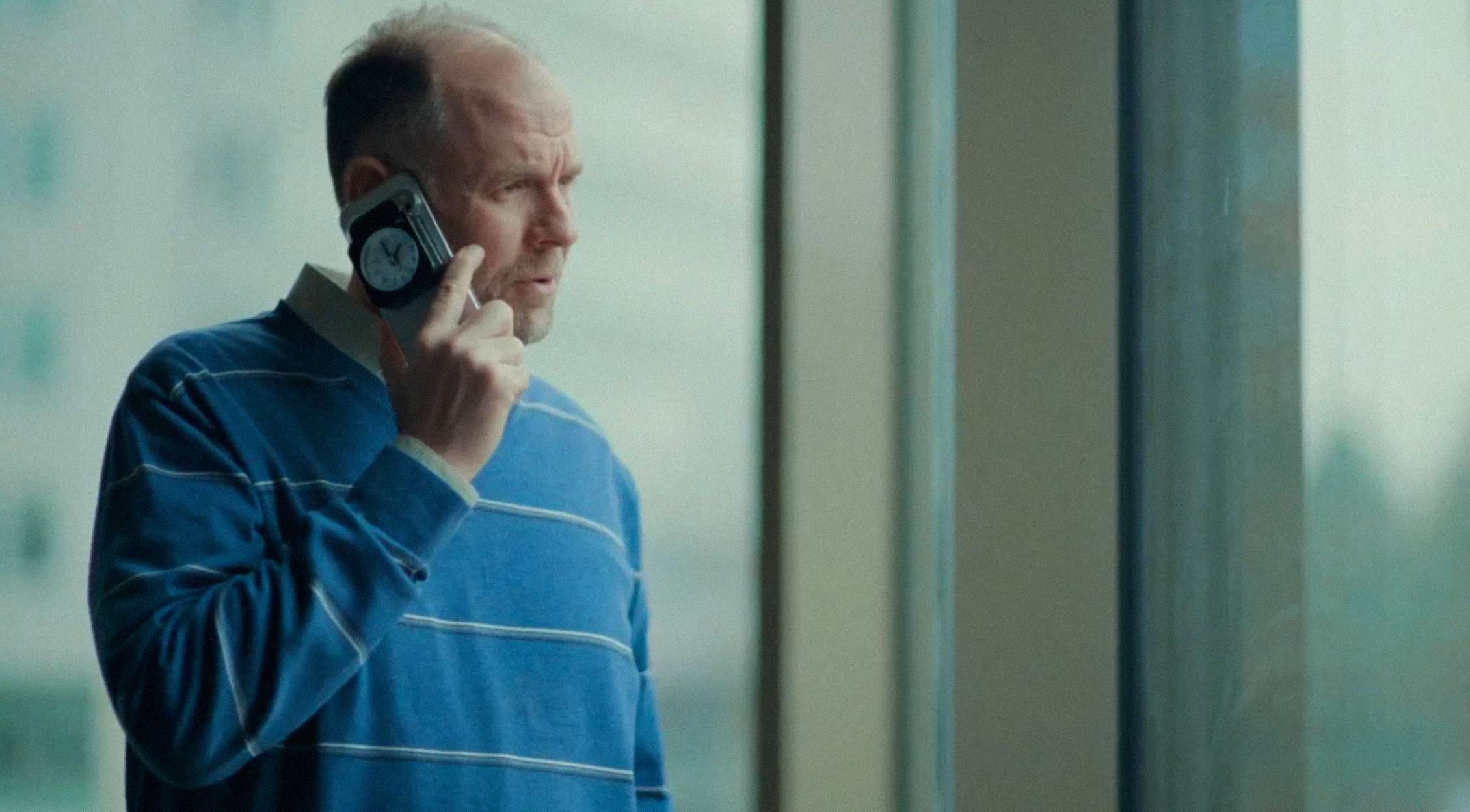 A man in a blue striped sweater talks on a cell phone with a small clock on its back, looking out a window.