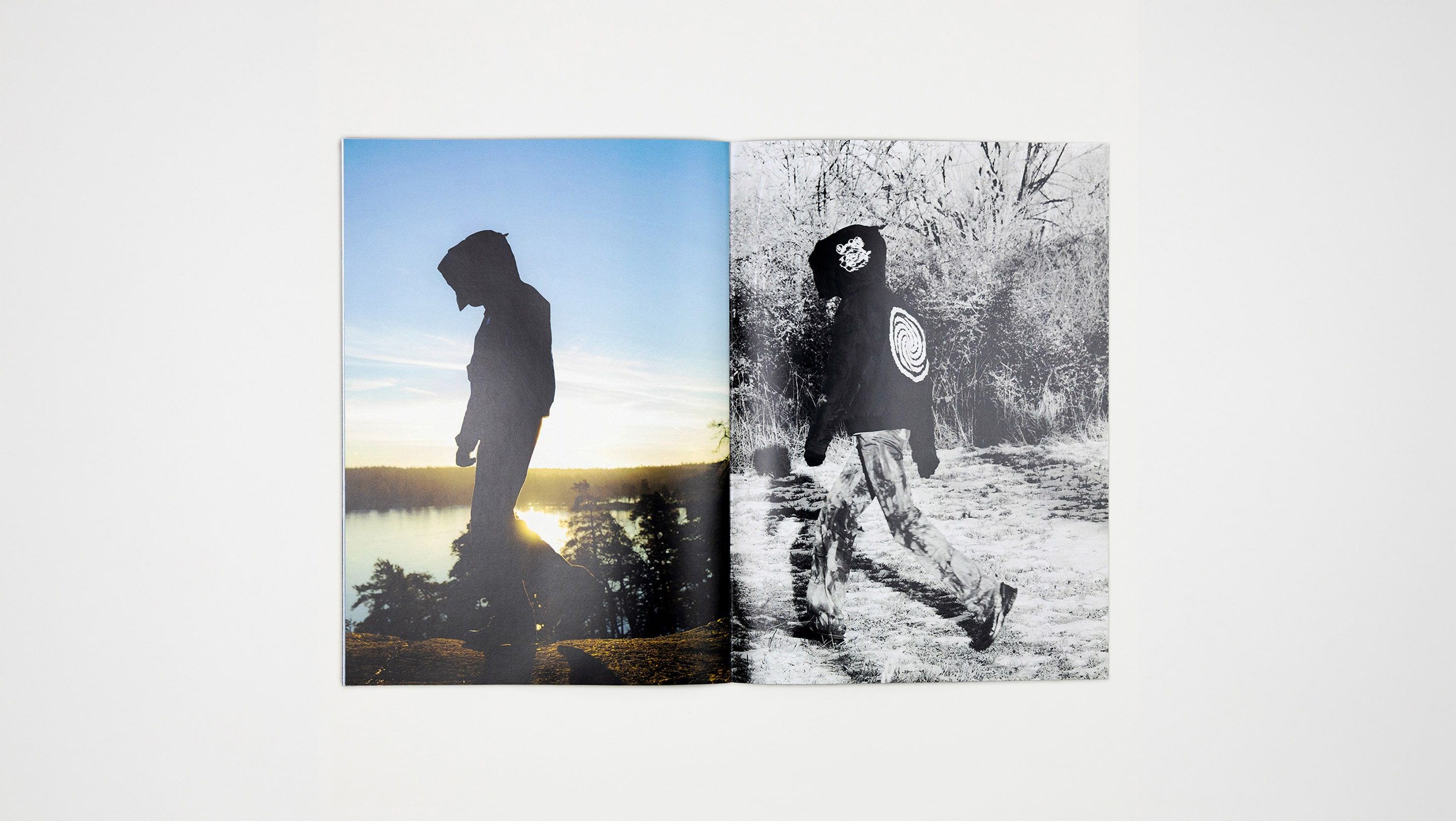 Flat-lay of a two-page zine spread: a colour sunset silhouette of a figure in the collection walking a coastal cliff on the left, and a black-and-white photo of the same figure walking through frosted brush wearing a hood with a hand-drawn spiral graphic on the back, on the right