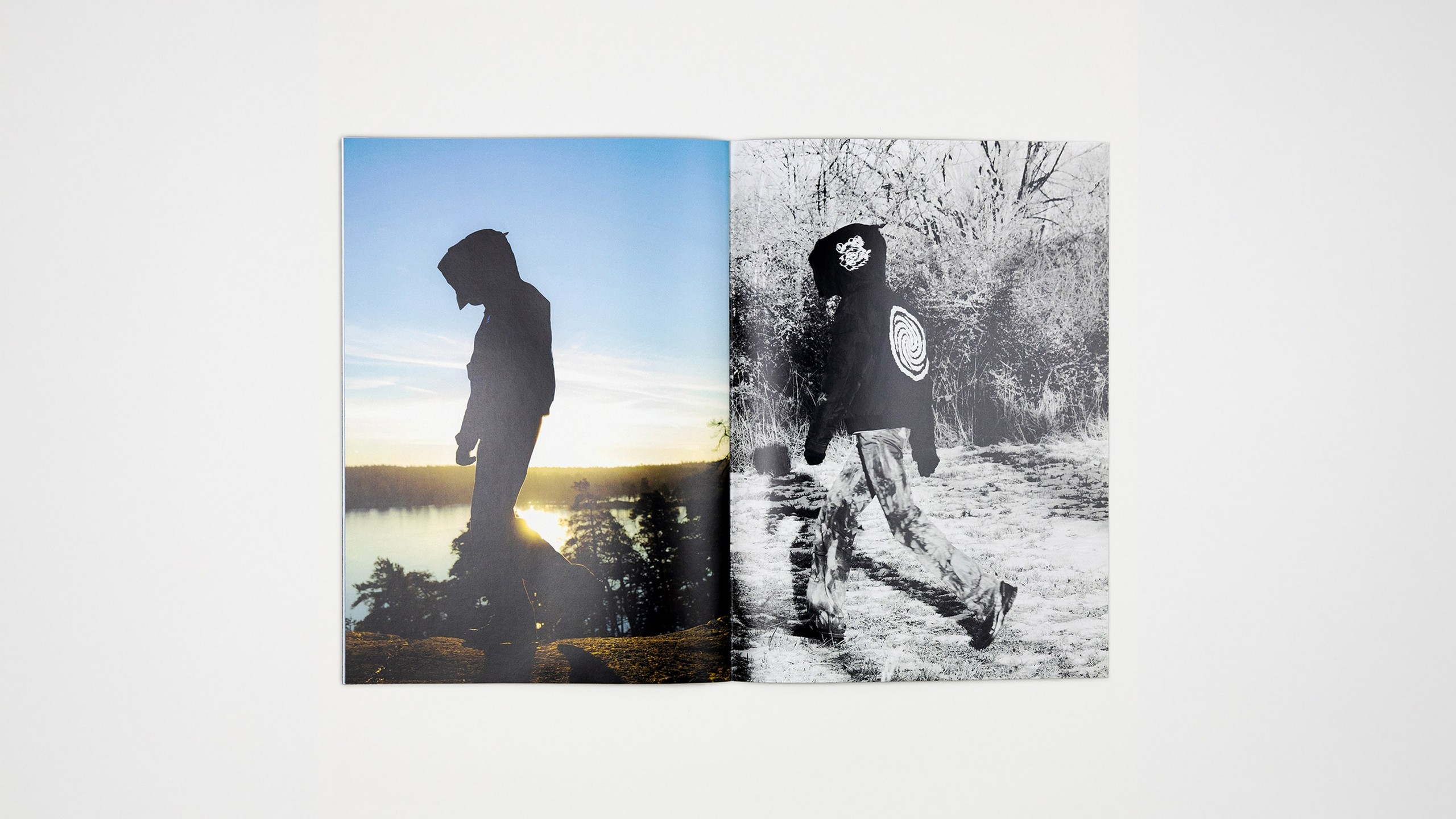 Flat-lay of a two-page zine spread: a colour sunset silhouette of a figure in the collection walking a coastal cliff on the left, and a black-and-white photo of the same figure walking through frosted brush wearing a hood with a hand-drawn spiral graphic on the back, on the right