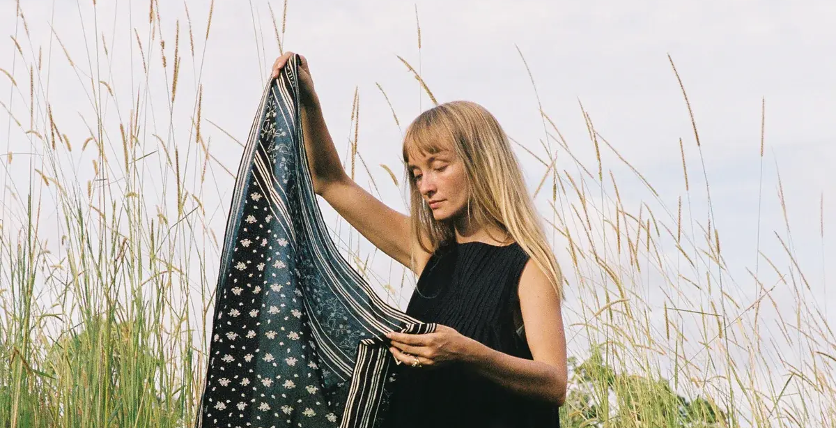 En kvinne med langt, blondt hår og svart singlet holder opp et mønstret skjerf med blomster- og stripemotiver, stående i et høyt gressfelt under en lys, overskyet himmel.