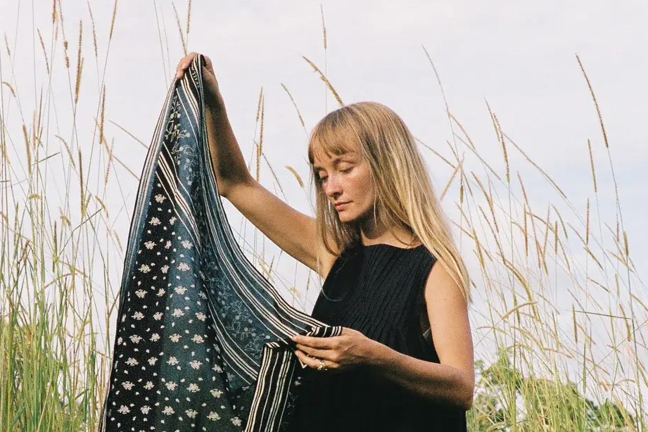 En kvinne med langt, blondt hår og svart singlet holder opp et mønstret skjerf med blomster- og stripemotiver, stående i et høyt gressfelt under en lys, overskyet himmel.
