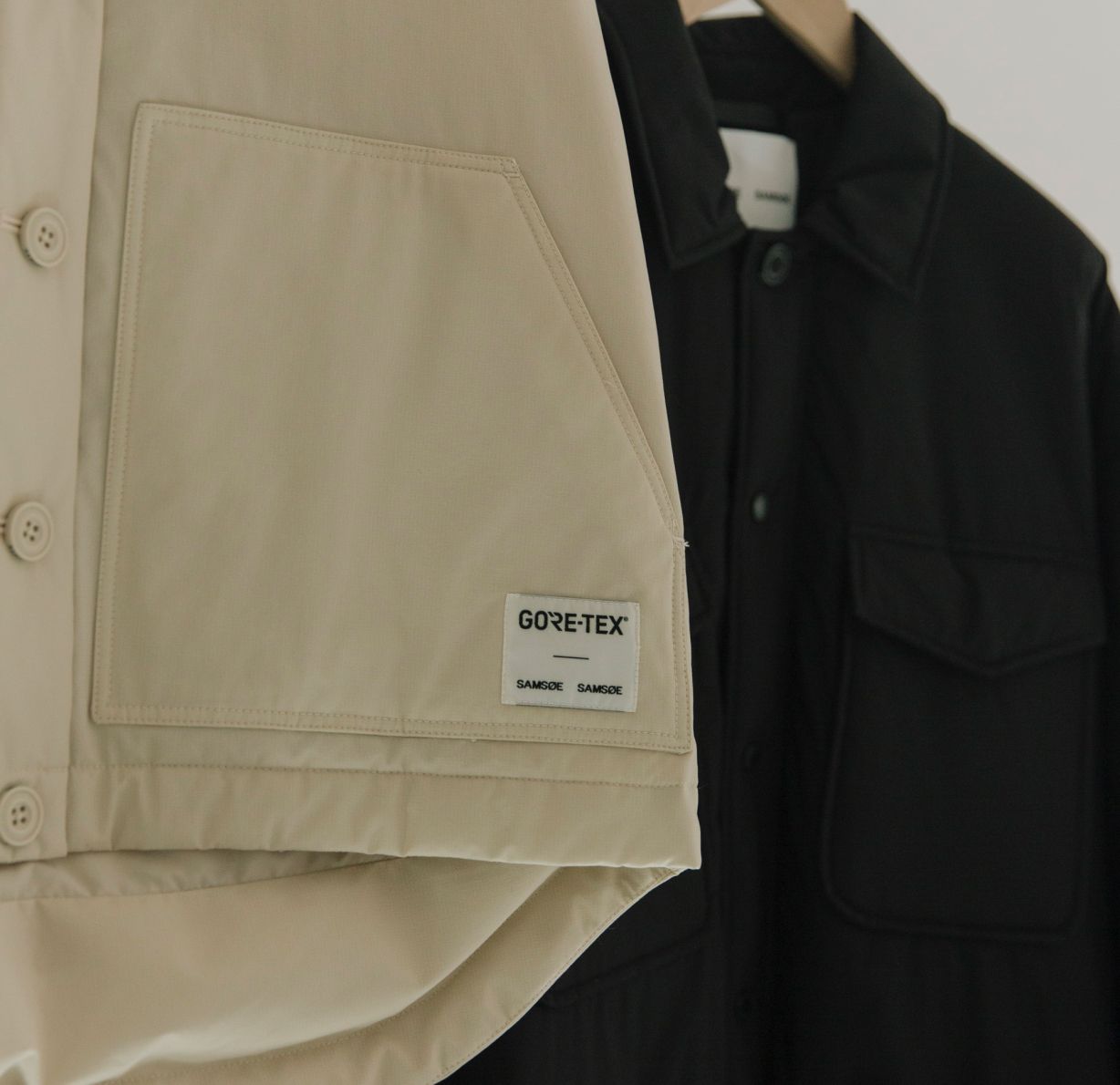 A woven GORE-TEX x Samsøe Samsøe label stitched onto a garment pocket, with two capsule jackets hanging behind it.