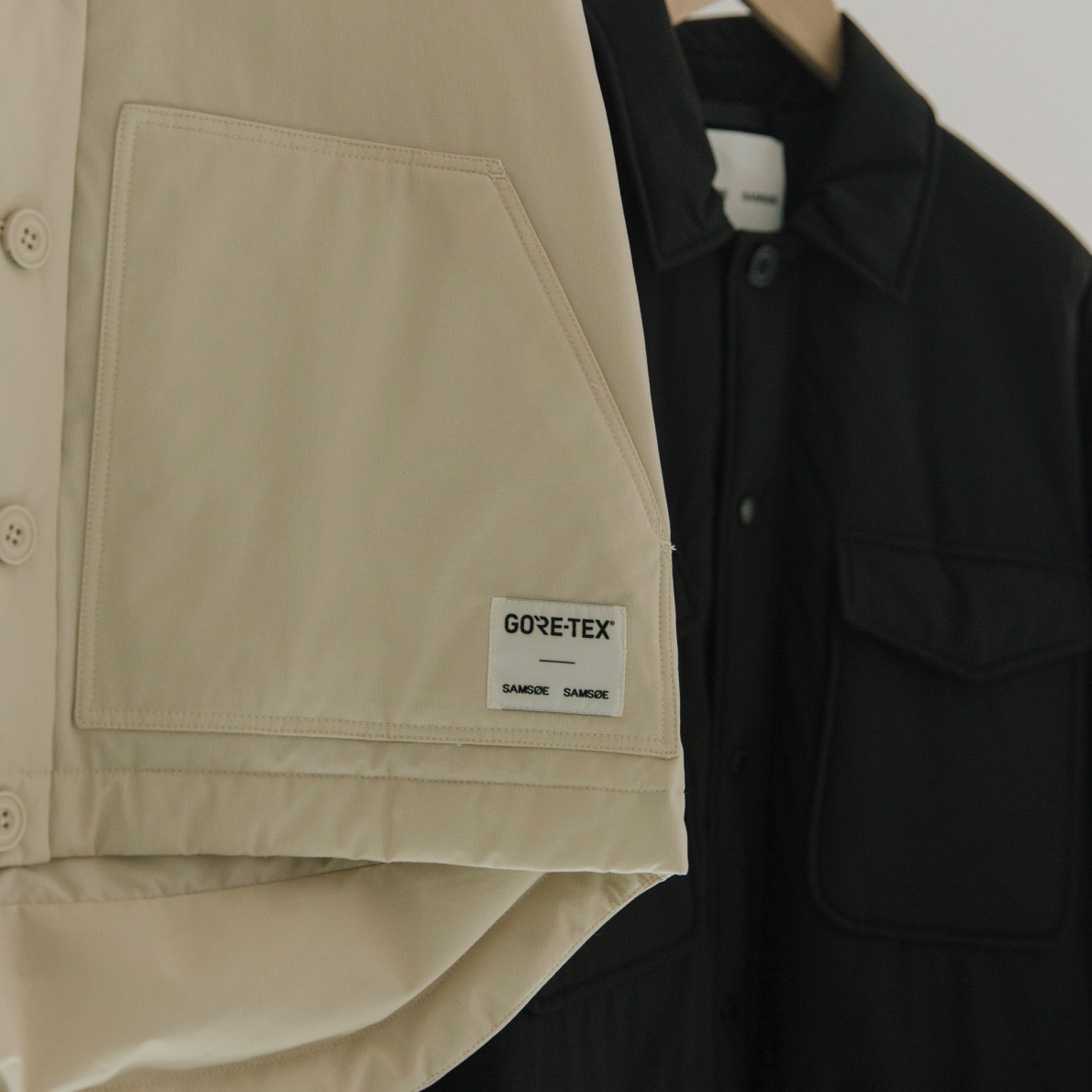 A woven GORE-TEX x Samsøe Samsøe label stitched onto a garment pocket, with two capsule jackets hanging behind it.