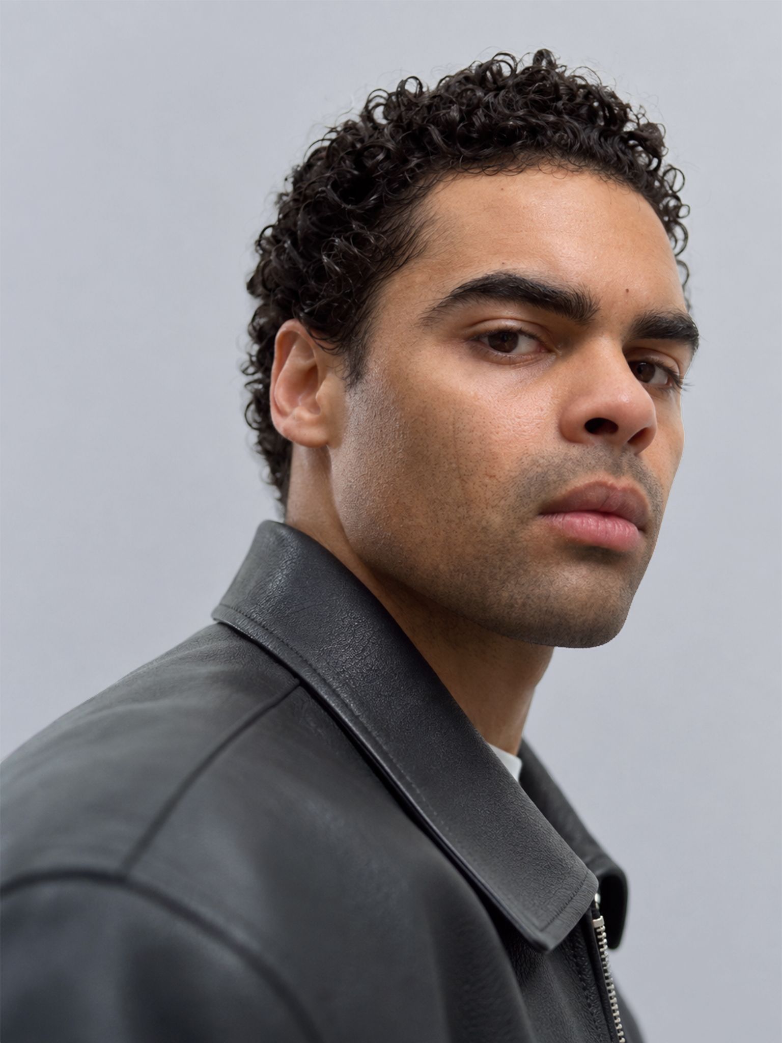 A man with dark curly hair wearing a black leather jacket looks directly forward.