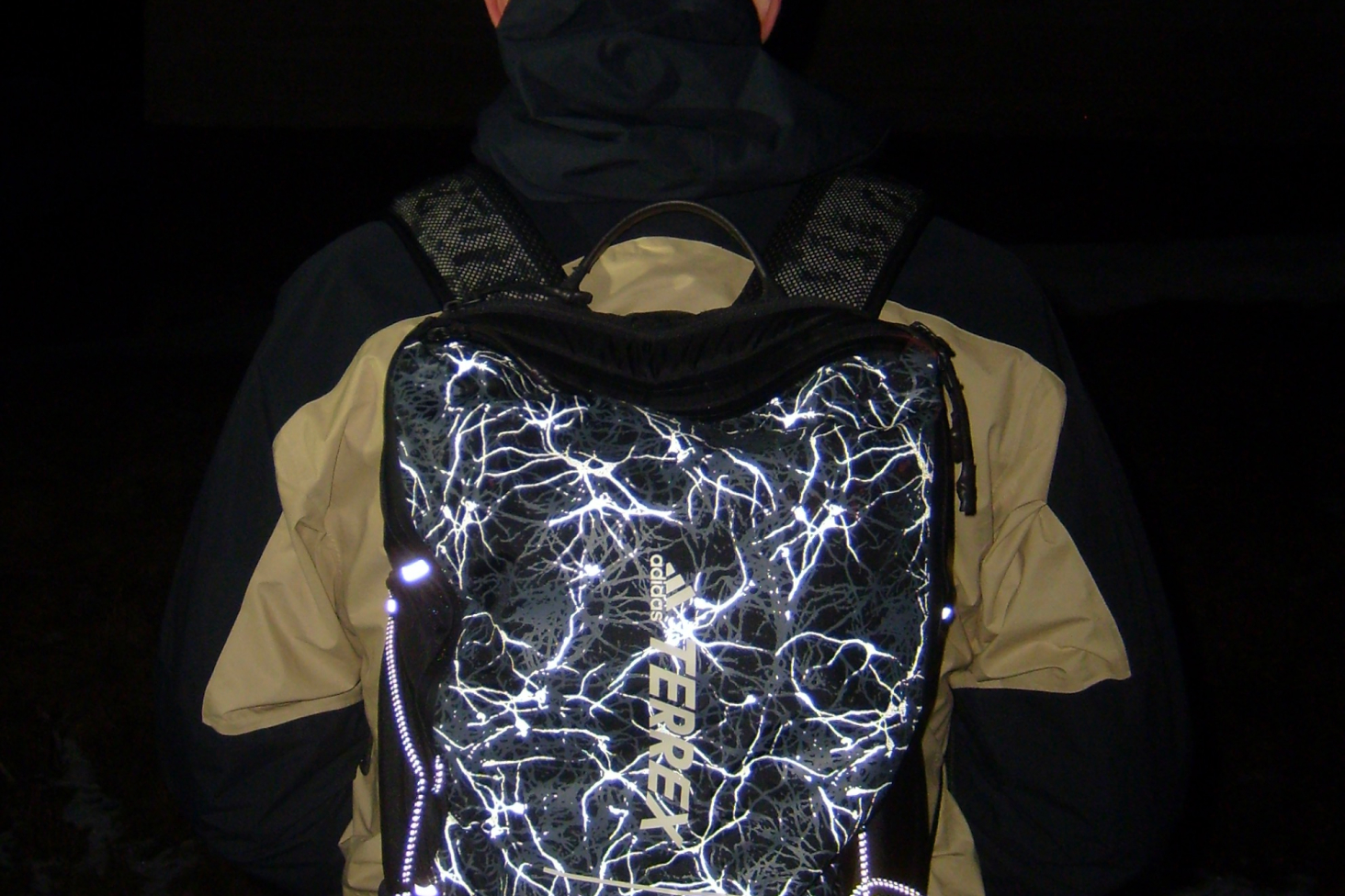 Back of a person wearing a reflective black backpack with a bright white lightning pattern against a dark background.