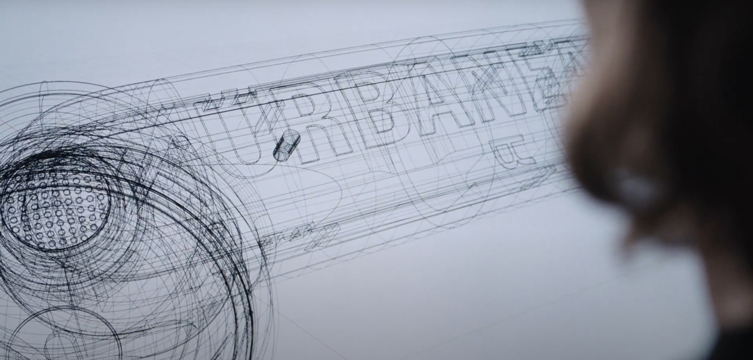 Wireframe CAD drawing of a cylindrical design with "URBANE" text and a perforated end, viewed by a person.