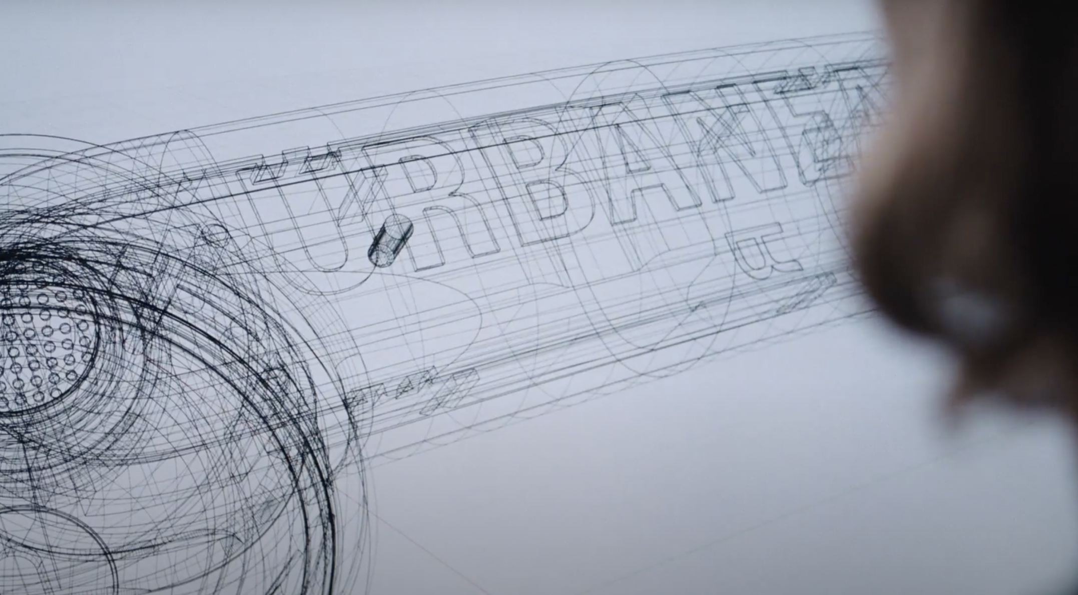 Wireframe CAD drawing of a cylindrical design with "URBANE" text and a perforated end, viewed by a person.