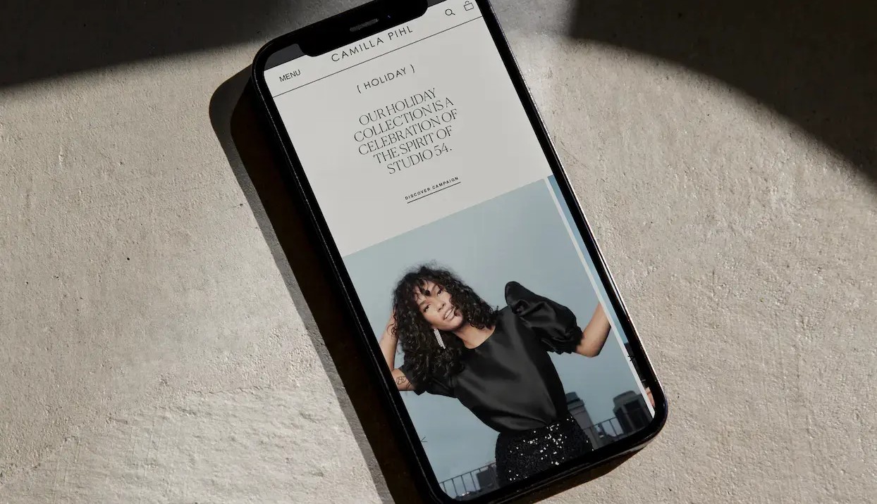 Black smartphone displaying a fashion website with a woman in a black satin top and sequined pants, and "Holiday Collection" text.