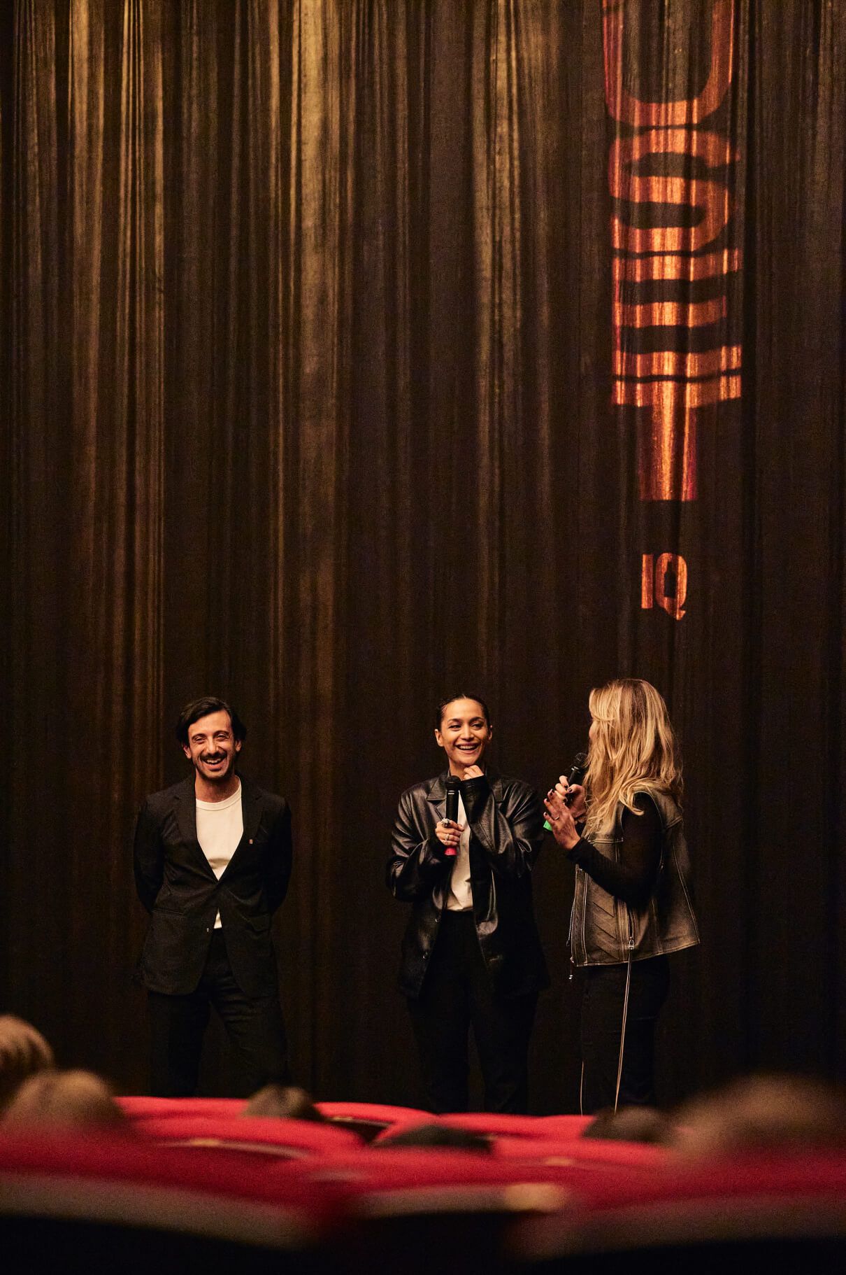 Three women on a stage holding microphones and laughing at the Ruset premiere, in front of a dark curtain lit with an orange RUSET title and the IQ mark.