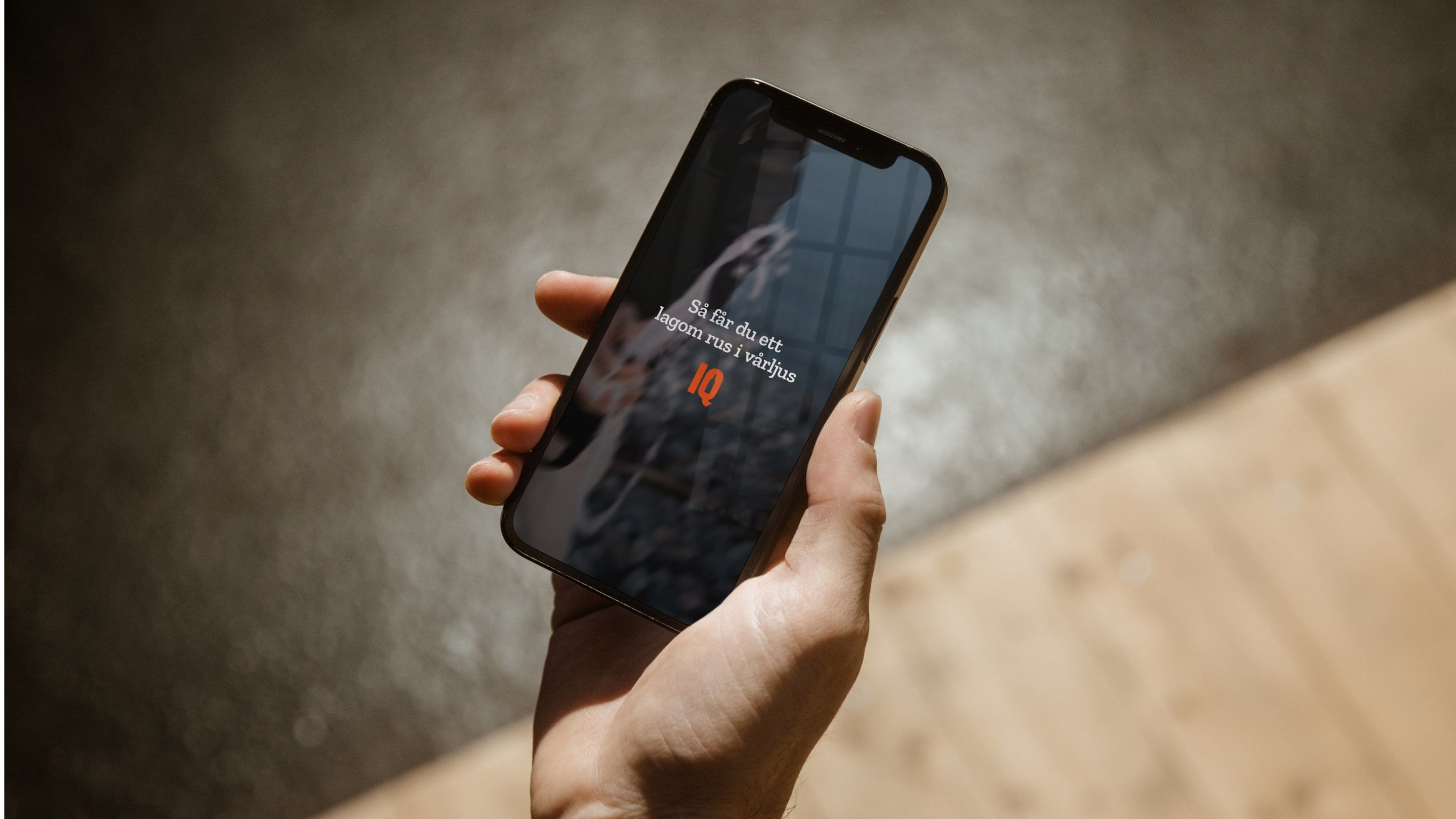 A hand holding up a phone showing a dark video still captioned "How to get a moderate buzz in spring light" with the IQ wordmark, against a blurred concrete and wood backdrop.