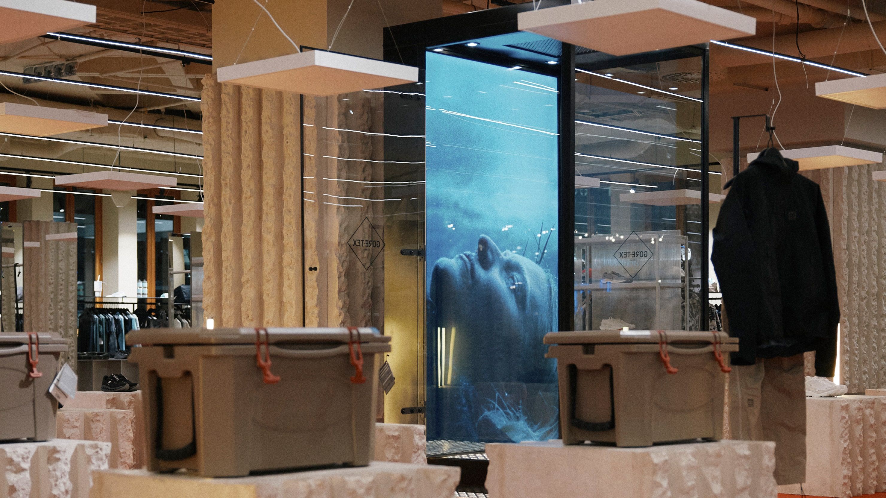 A man in a 66°North GORE-TEX jacket and pants standing inside a vertical glass column as rain pours over him, a projected ocean backdrop behind, the GORE-TEX diamond logo etched at bottom right.