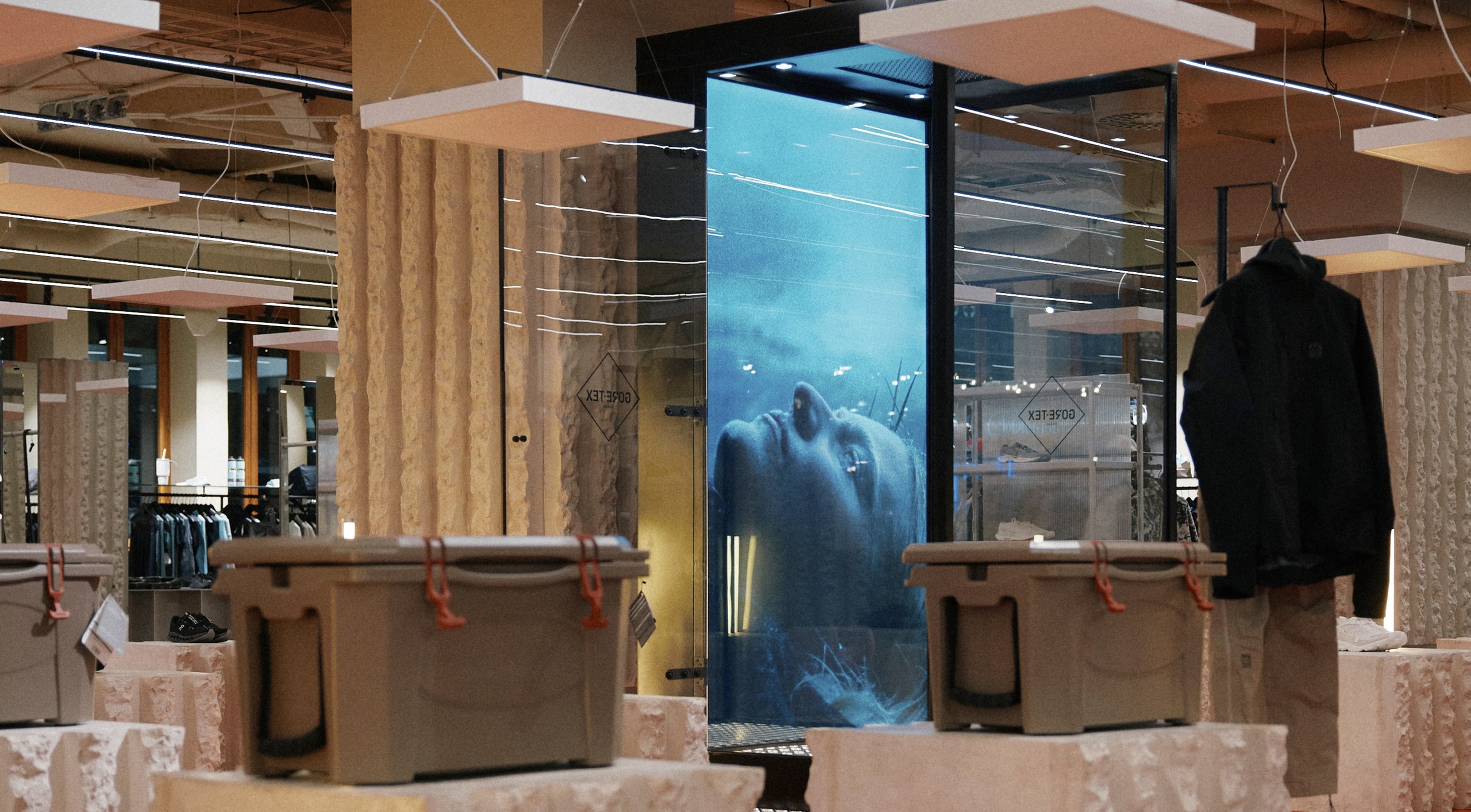 A man in a 66°North GORE-TEX jacket and pants standing inside a vertical glass column as rain pours over him, a projected ocean backdrop behind, the GORE-TEX diamond logo etched at bottom right.