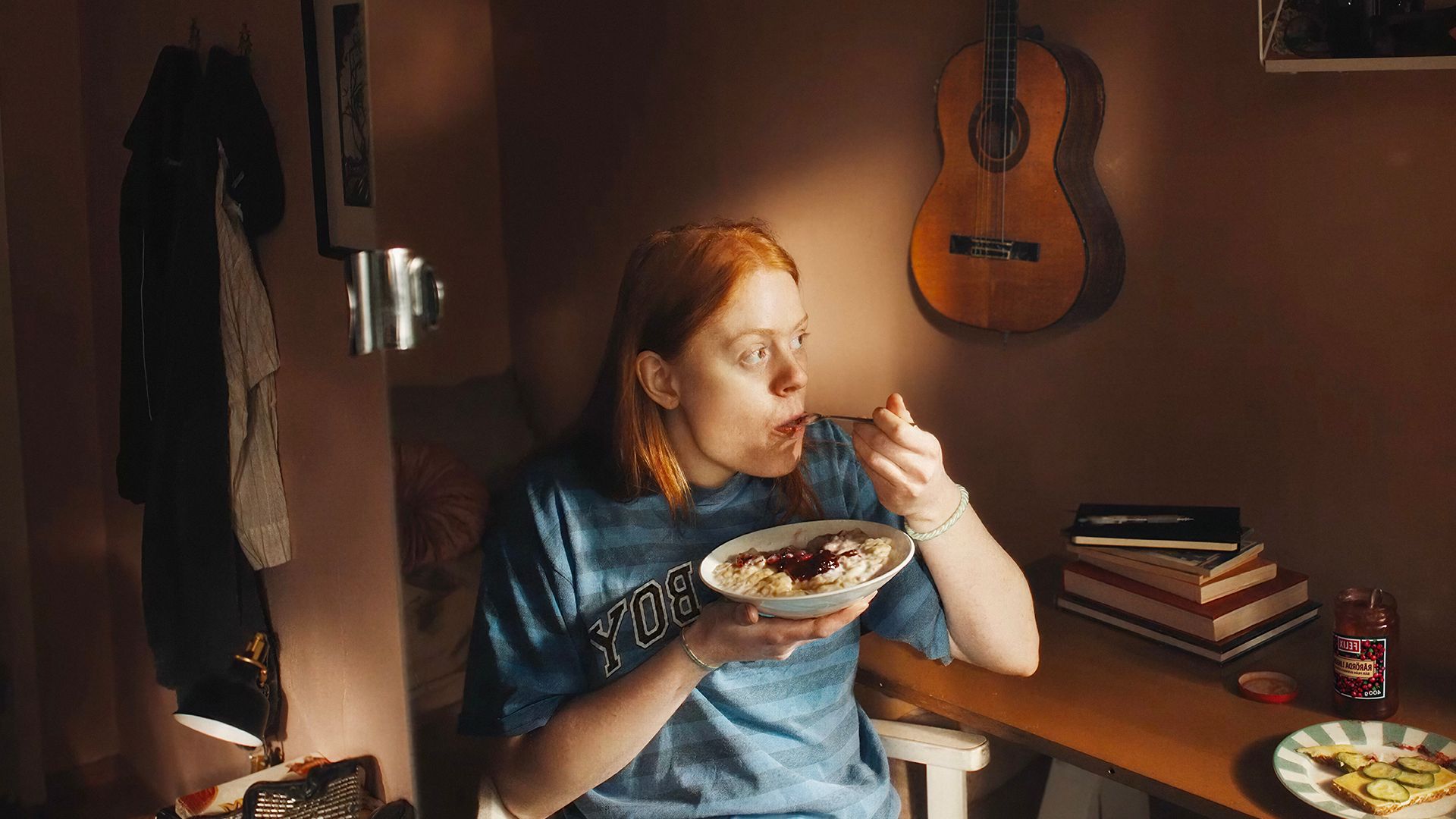 A red-haired woman eats oatmeal from a bowl at a sunlit desk with a guitar on the wall.