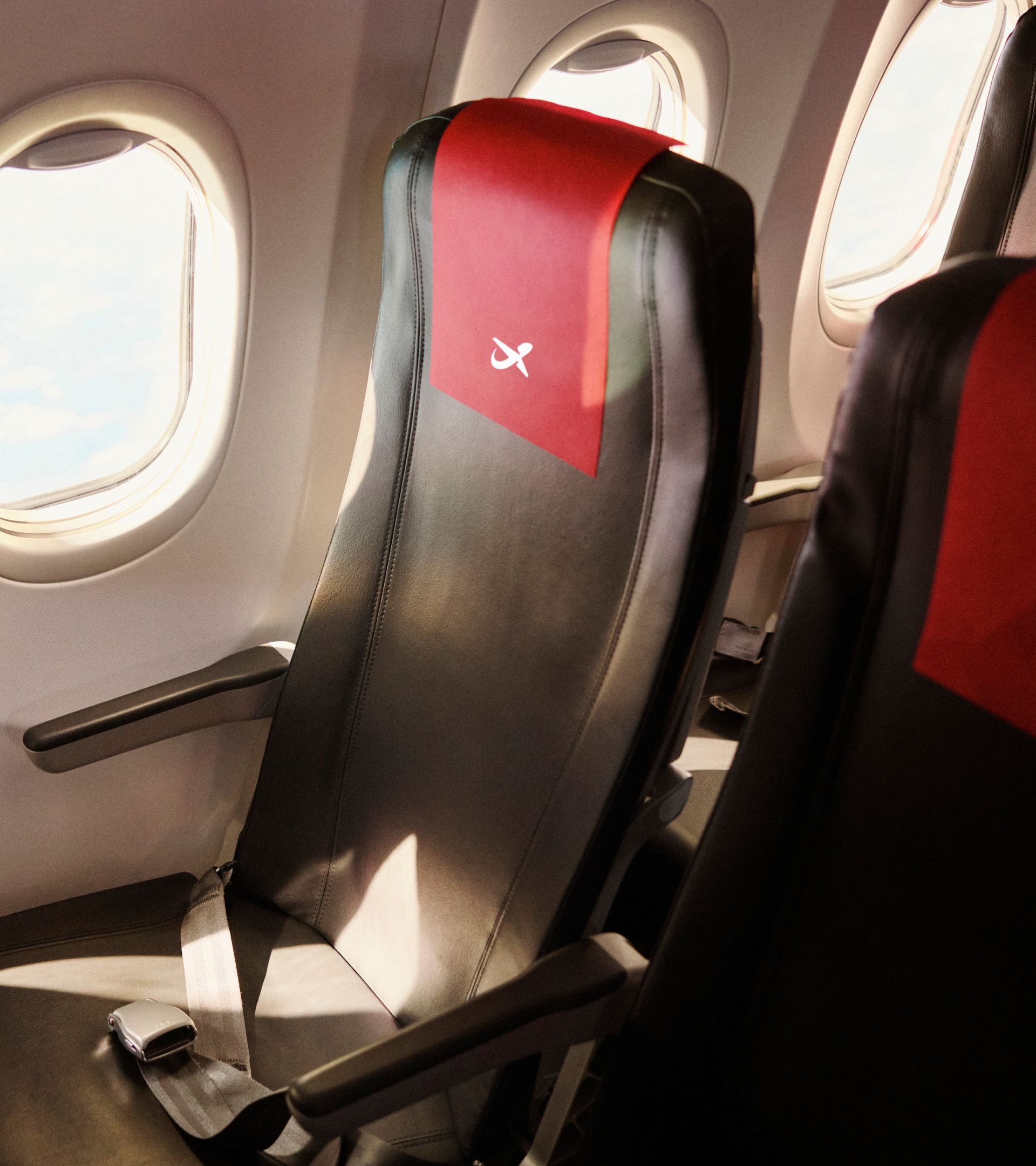 An aircraft seat with a red headrest cover carrying the Norwegian symbol, beside a cabin window