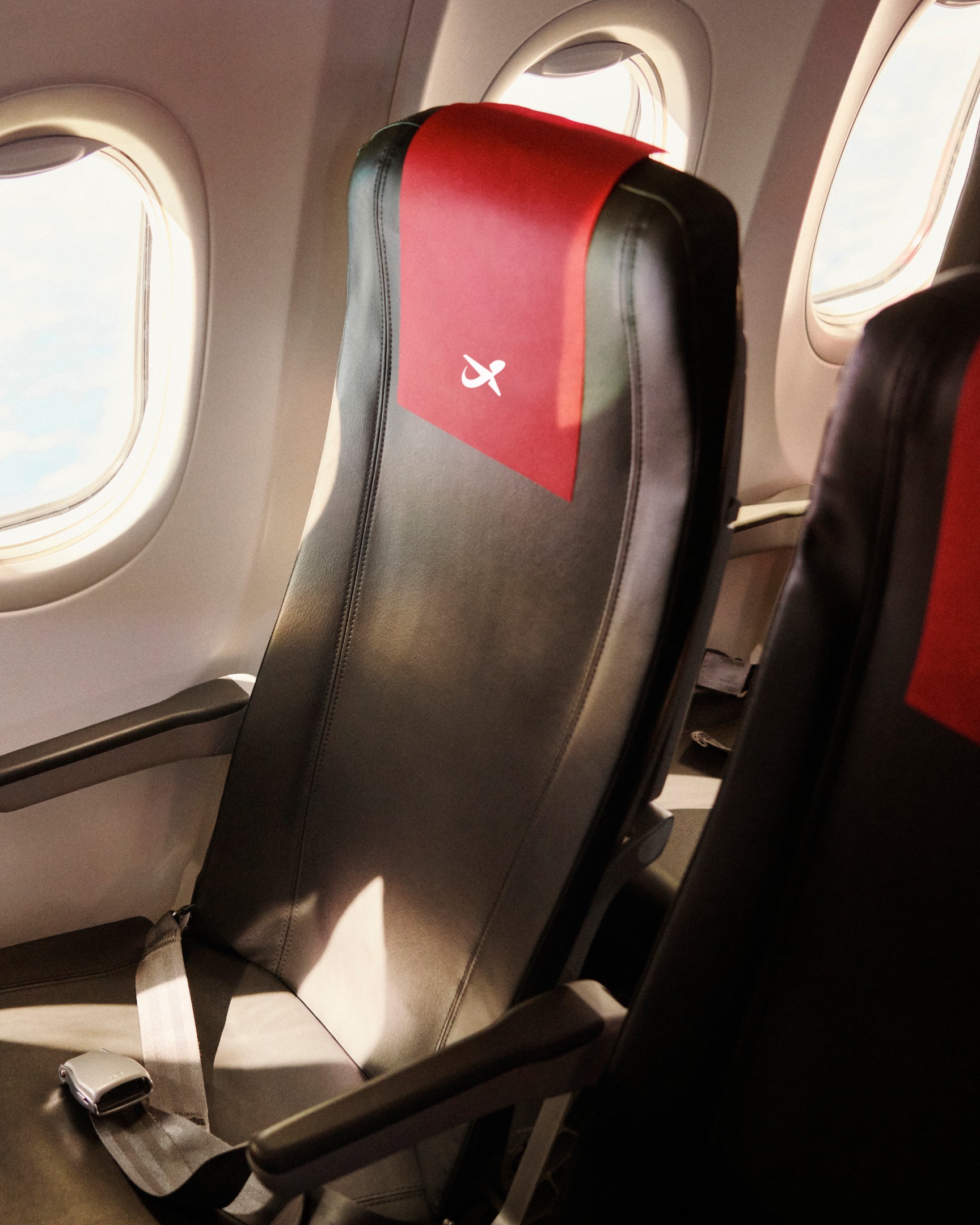 An aircraft seat with a red headrest cover carrying the Norwegian symbol, beside a cabin window