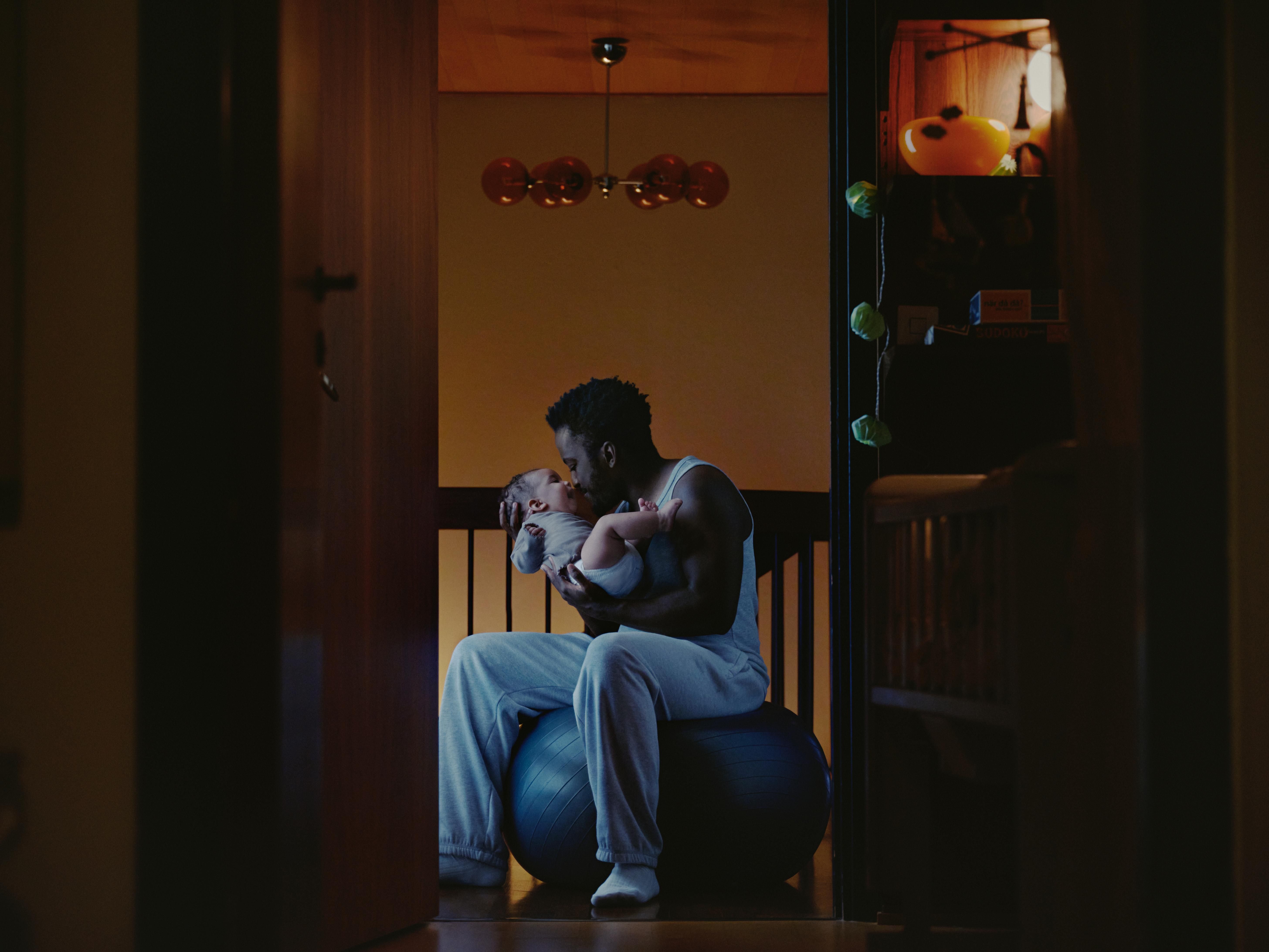 A Black man tenderly holds a baby, looking at it, while sitting on an exercise ball.