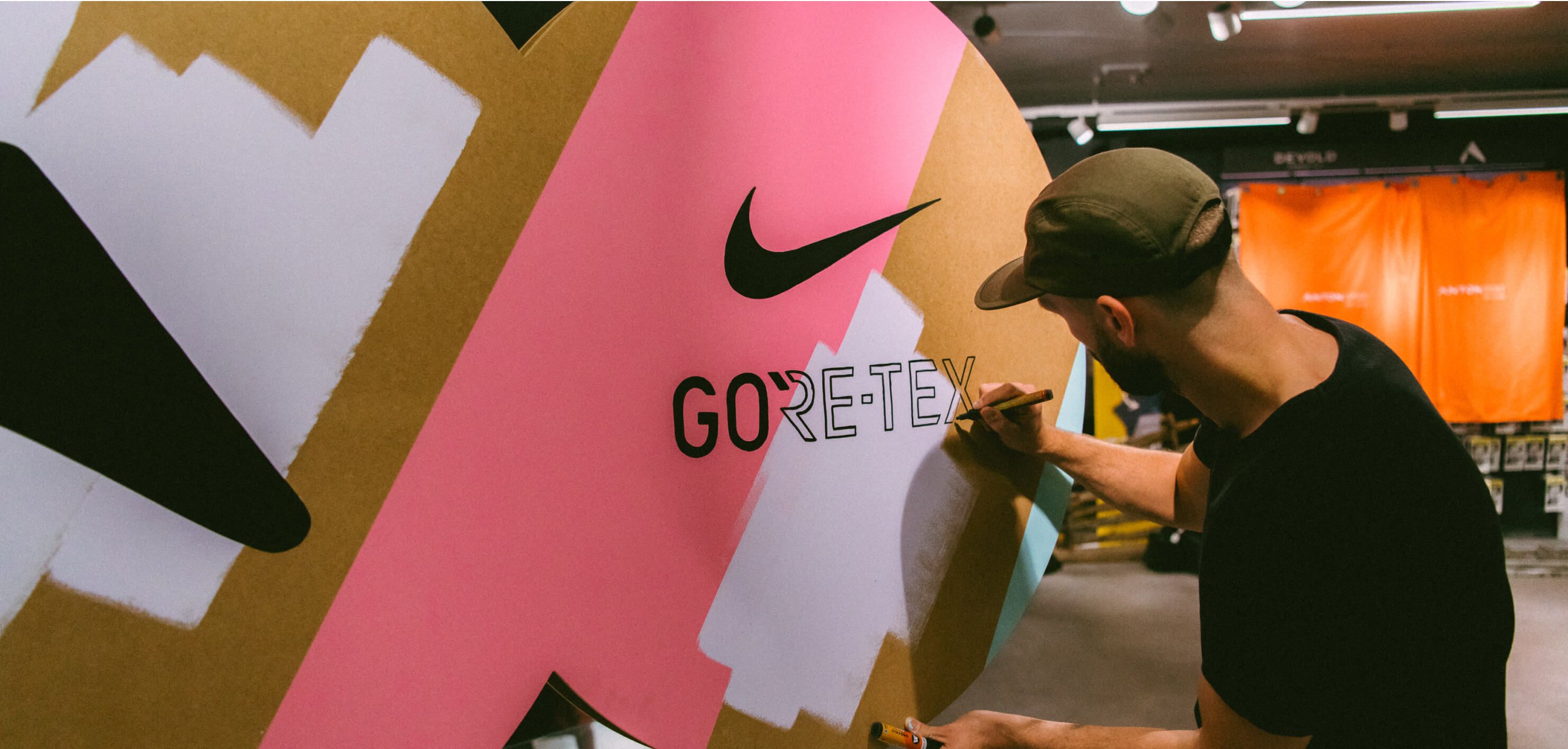 An artist mid brush stroke, painting a Nike swoosh and a GORE-TEX wordmark onto a giant sneaker shaped cardboard installation, in pink, gold and white paint, at the GORE-TEX x Nike Pegasus launch event in Oslo.