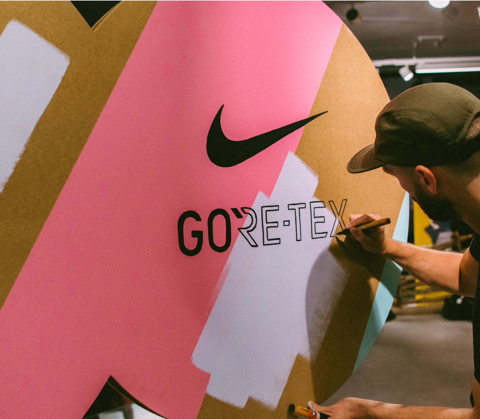 An artist mid brush stroke, painting a Nike swoosh and a GORE-TEX wordmark onto a giant sneaker shaped cardboard installation, in pink, gold and white paint, at the GORE-TEX x Nike Pegasus launch event in Oslo.