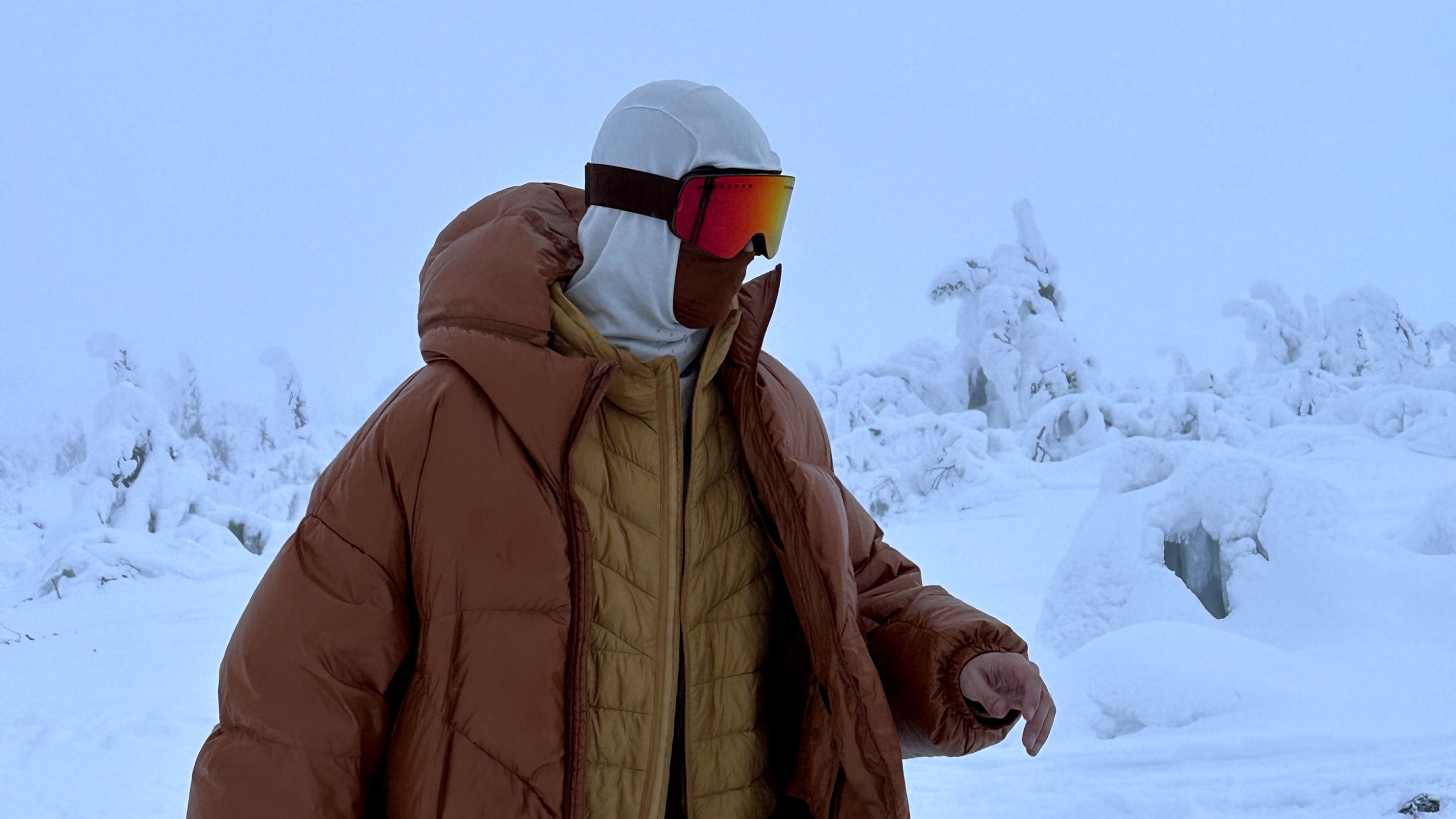 A figure wrapped in an oversized rust-orange puffer jacket, mirrored ski goggles reflecting orange light, snow-laden trees behind.