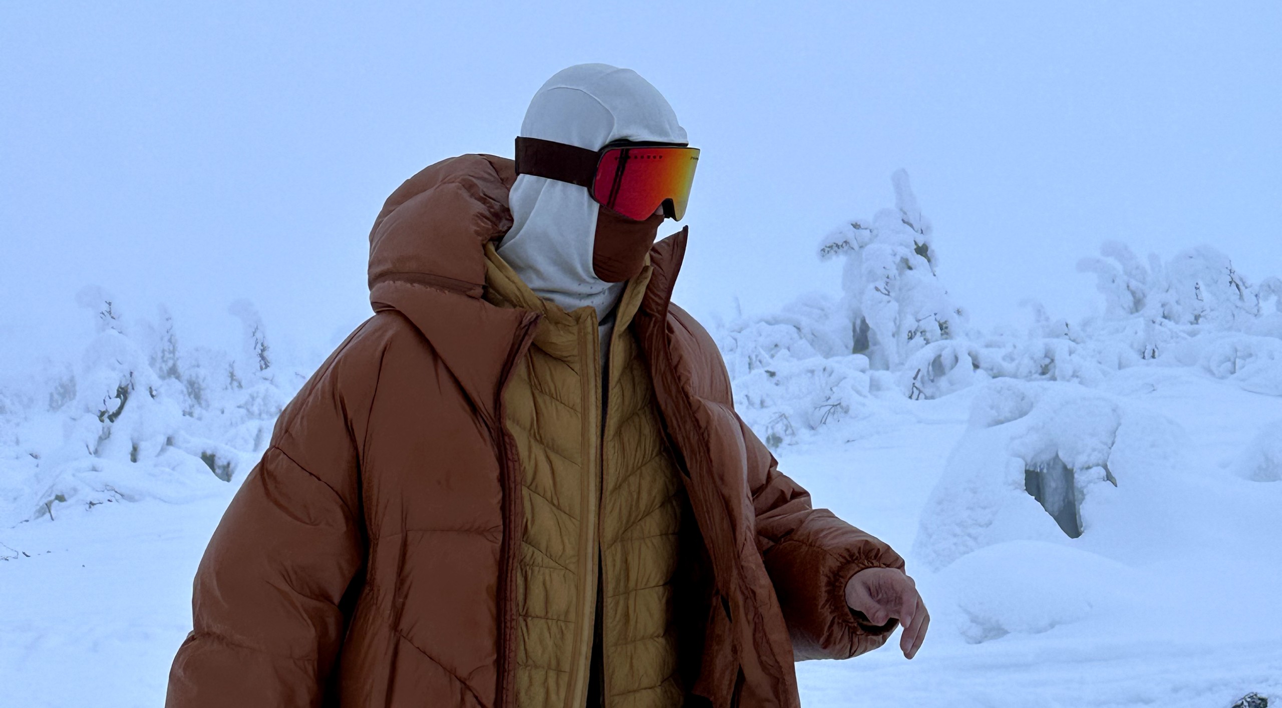 A figure wrapped in an oversized rust-orange puffer jacket, mirrored ski goggles reflecting orange light, snow-laden trees behind.