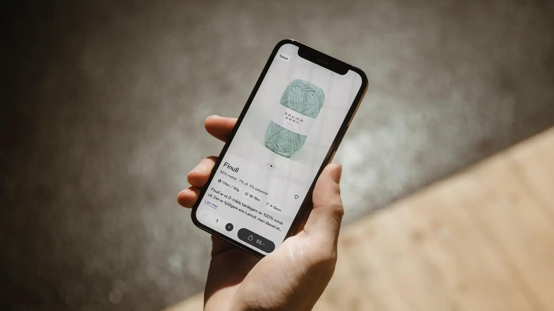 En hånd holder en mobil åpen på Rauma Garns nettbutikk, på produktsiden for garnet «Finull», som viser pris og en «legg i kurv»-knapp.