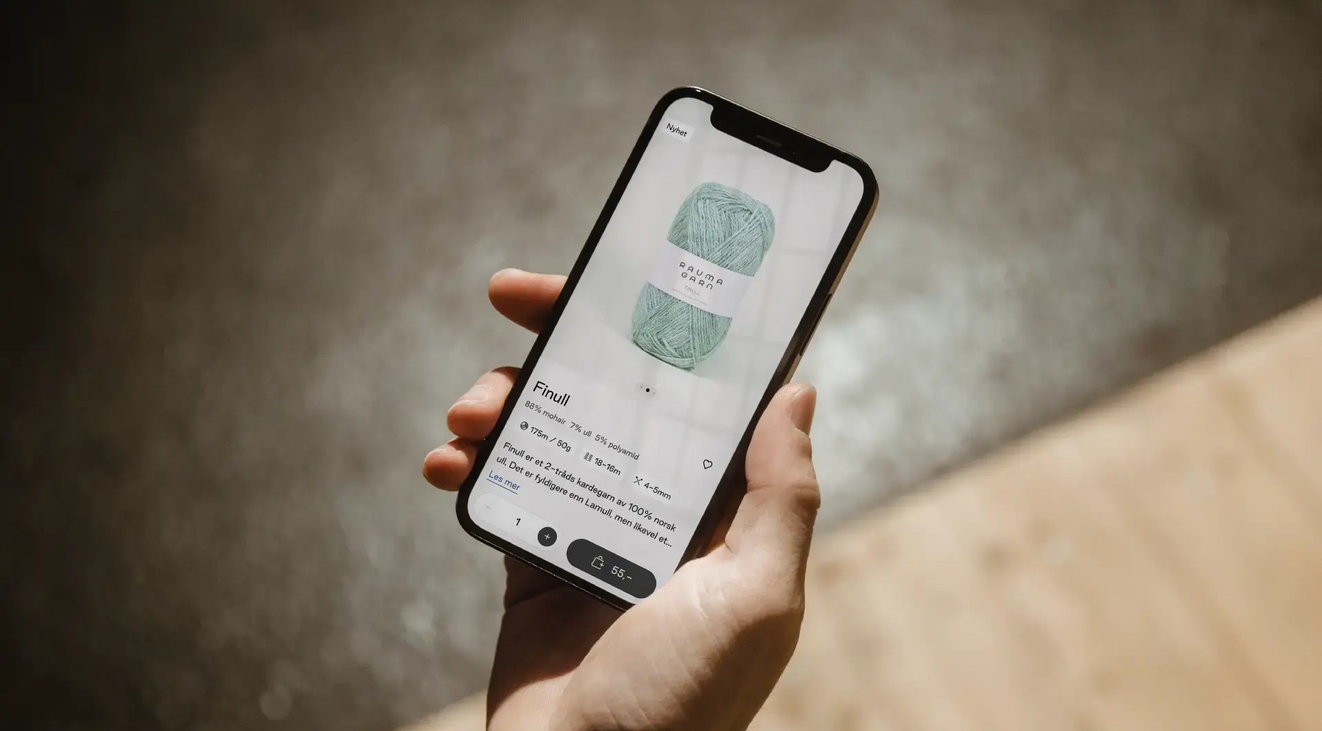 En hånd holder en mobil åpen på Rauma Garns nettbutikk, på produktsiden for garnet «Finull», som viser pris og en «legg i kurv»-knapp.
