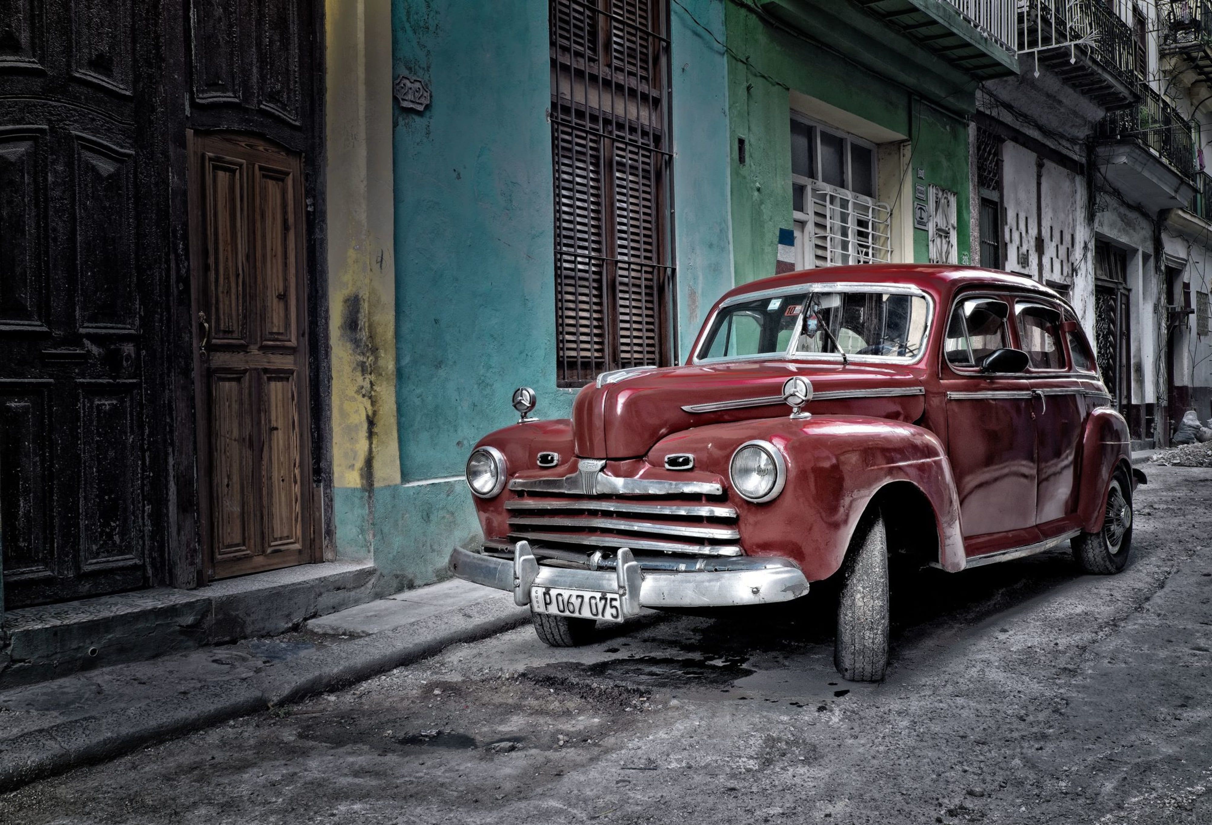 A dark red 1940s American car parked on a narrow Havana street, its chrome grille catching the light against faded pastel walls.