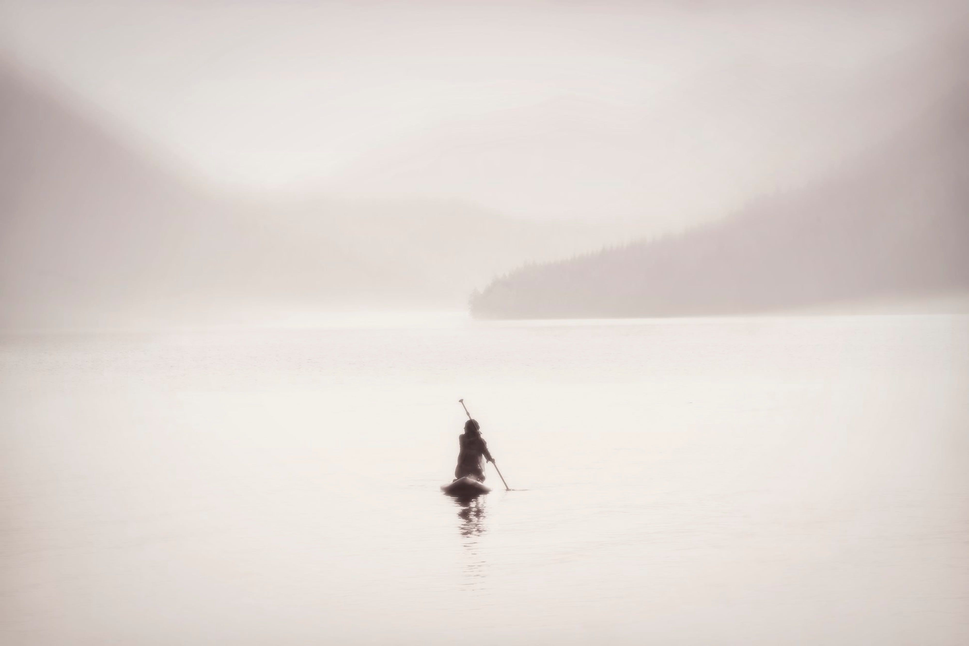 A lone figure stands waist deep in still pale water beneath a hazy headland, the horizon almost dissolved in mist.
