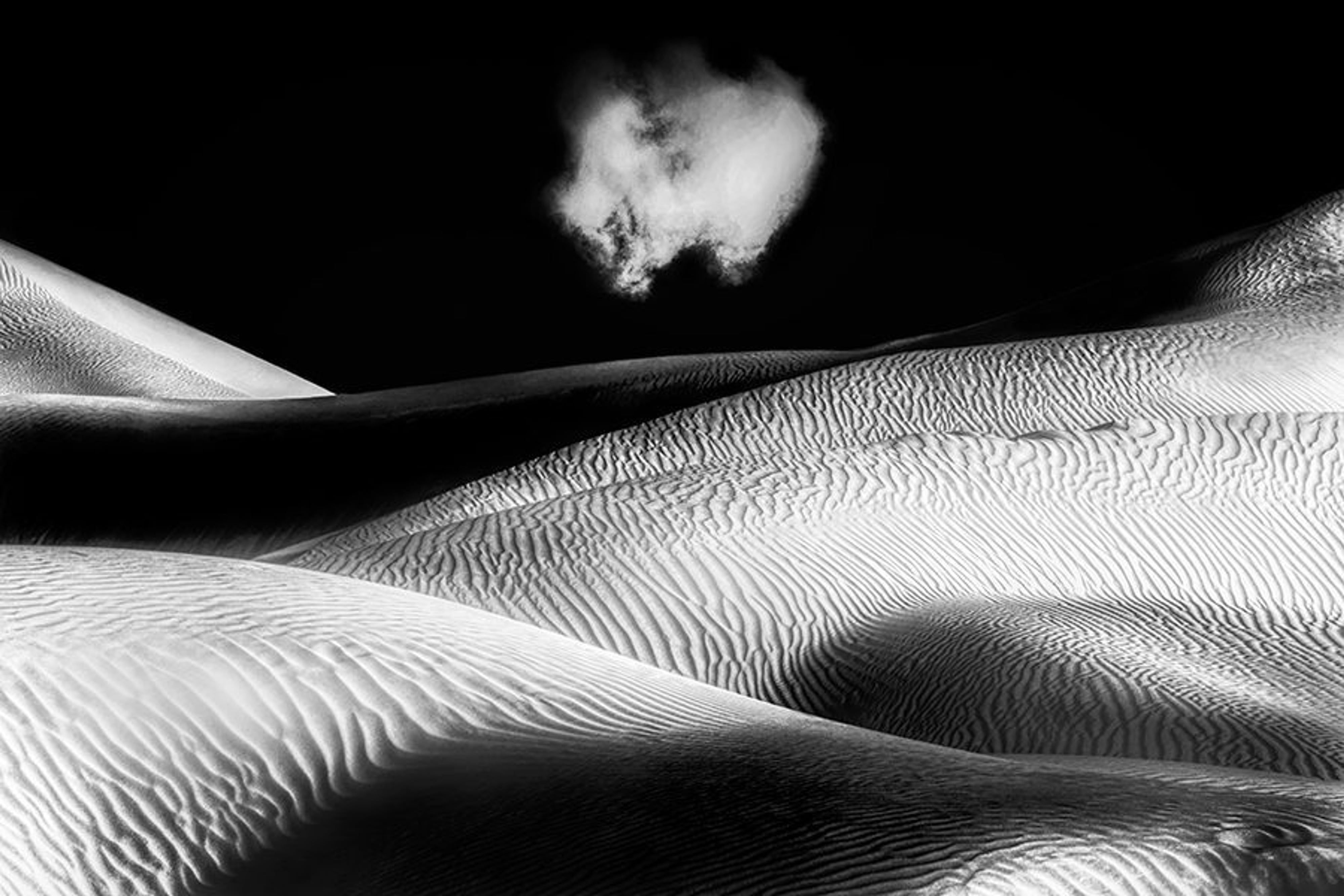 A single small cloud hangs above a rippled sand dune, its lit crest curving across a black sky.