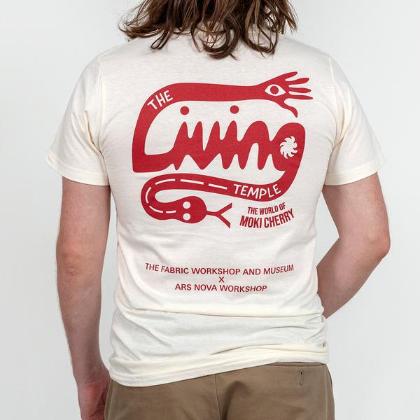 A model turned away from the viewer wears an off-white t-shirt. The back of the shirt features a red graphic logo that reads "The Living Temple: The World of Moki Cherry". The tail of the 'L' ends in a hand with an eye while the tail of the 'G' ends in a snake with a forked tongue protruding. The lower portion of the shirt reads "The Fabric Workshop and Museum x Ars Nova Workshop"