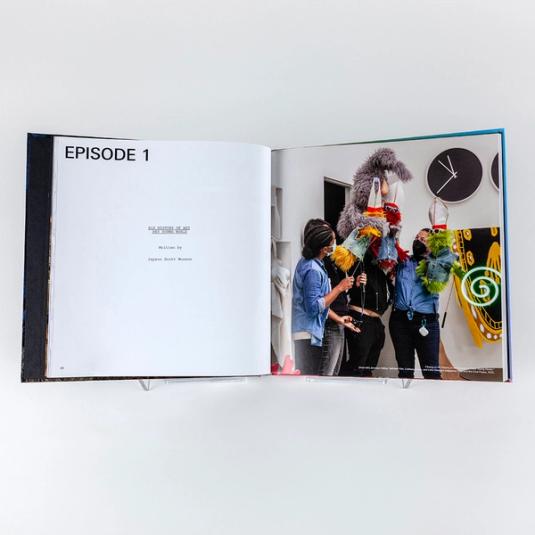 A book spread featuring one page of text on the left that reads "Episode 1: His History of Art Hey Young World, Written by Jayson Scott Musson. On the right is a behind-the-scenes photograph of four puppeteers animating three, smaller colorful (one yellow, red, and green) furry puppets in denim vests, sunglasses, and pope hats as well as one larger, scraggly gray rabbit puppet. They are on the set of filming for Jayson Musson: His History of Art.