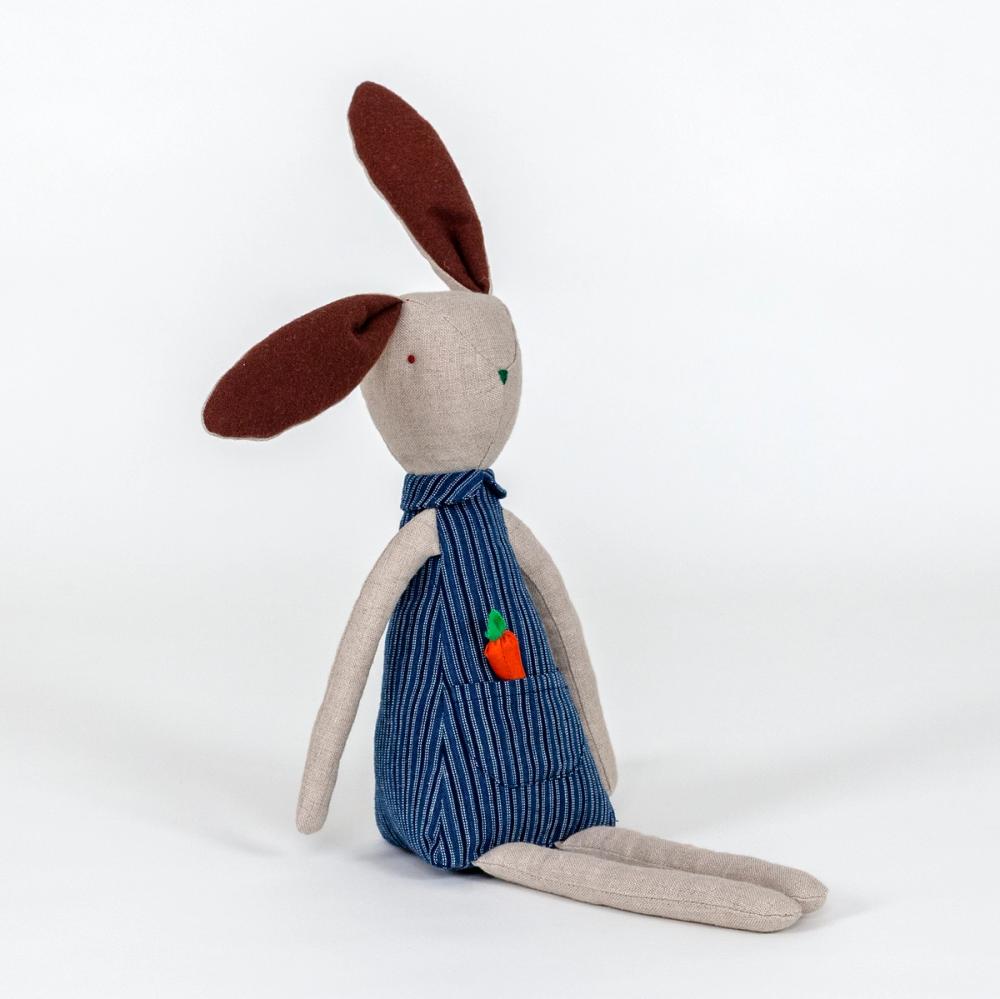 A photograph of a plush rabbit made of light beige woven fabric characterized to have a pointy nose, triangular shaped body, and long thin arms and legs. The body of the rabbit is made of a primarily blue patterned fabric and includes a pocket with a removable plush orange carrot.