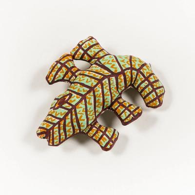 A photograph of a small plush toy resembling an alligator, printed in light green with dark brown lines and V-shaped orange scales. Its blunt legs splay out from either side of its body. Its eye is long, orange, and almond-shaped, outlined in dark brown with a pupil of the same color.