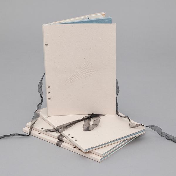 A photograph of a cream colored paperback book with eyelet binding and a sheer black ribbon attached to the cover. The material of the book appears to be a textured card stock with the title "Material Dreams" embossed on the front. One book is standing up on three of the same books.