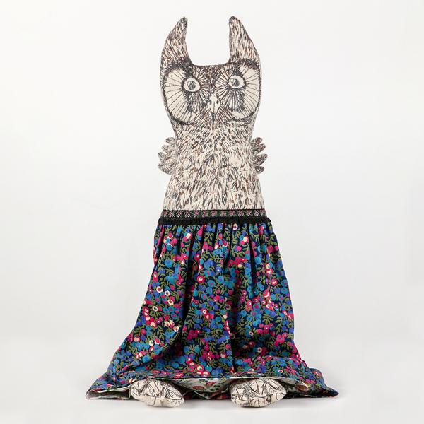 A photograph of a reversible flip doll. In this instance, a plush owl with drawn details stands upright with a skirt of florals screenprinted in cool colors. The owl has little feet peeking out from beneath the skirt's hem.