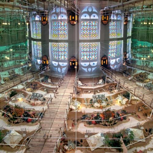 A detail of a book spread featuring an art installation consisting of a refracted architectural space ending with a repeated and mirrored stained glass window. The lower refractions are divided by wooden boardwalks with smaller clusters of sand, furniture, and assorted objects.