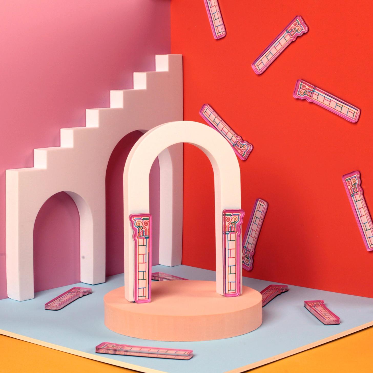 A colorful tabletop scene with a box-like set featuring pink, red, and blue walls and geometric arches. Clear plastic magnets shaped like classical columns, printed with white and multicolor decorative designs, are scattered across the scene and attached to the walls