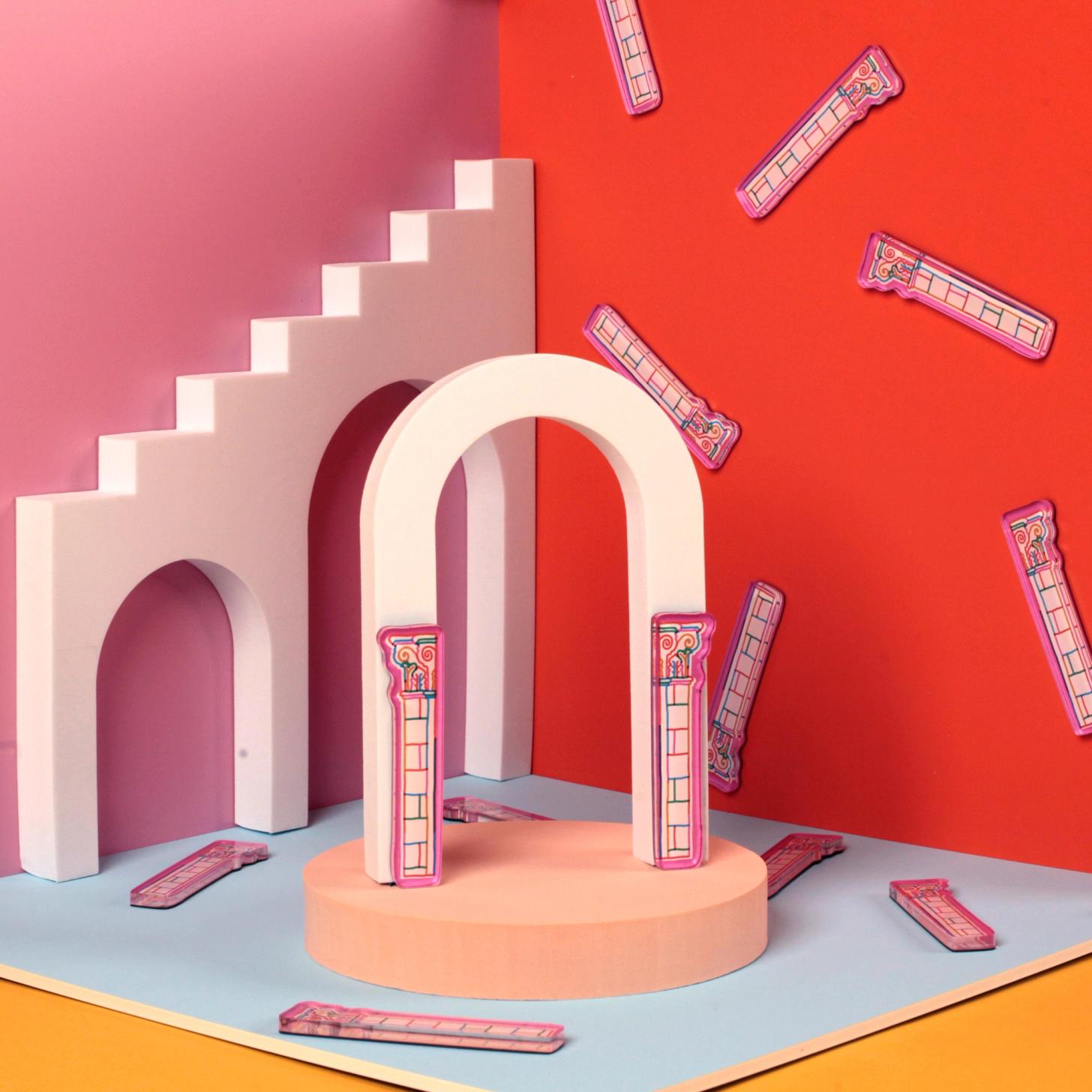 A colorful tabletop scene with a box-like set featuring pink, red, and blue walls and geometric arches. Clear plastic magnets shaped like classical columns, printed with white and multicolor decorative designs, are scattered across the scene and attached to the walls