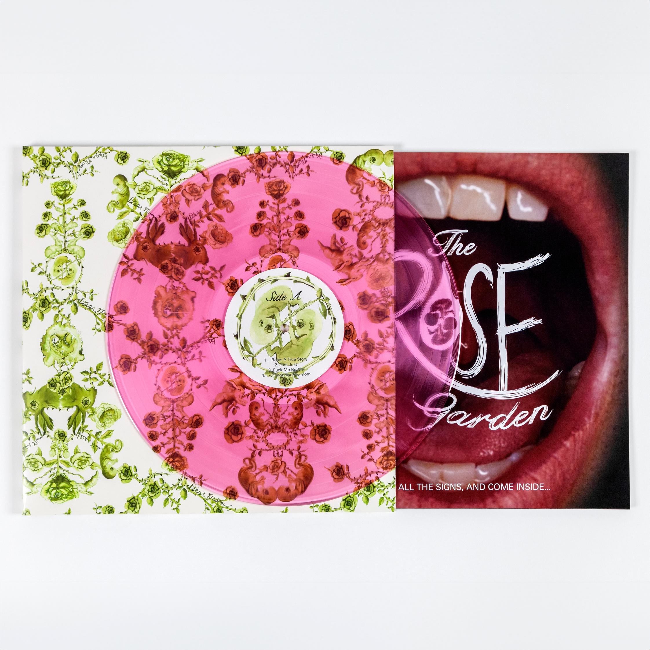 A vinyl record cover featuring a hand illustrated green toile-like pattern made up mostly of roses but also features twin fetuses and rabbits. This motif is on a solid cream background. Coming out of the cover to the right is a booklet featuring a close-up photo of a mouth open wide. The cover of the book reads "John Jarboe's The Rose Garden IGNORE ALL THE SIGNS, AND COME INSIDE..." Laying on top of both the record cover and booklet is a see-through pink vinyl.