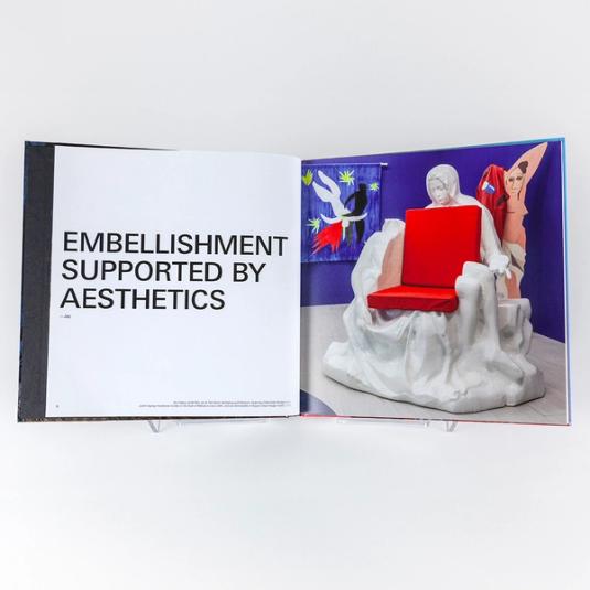 A book spread featuring one page of large text on the left that reads "Embellishment supported by aesthetics" and is quoted by "Jay" who is Jayson Musson's character in the exhibition, His History of Art. The page on the right is an installation photo from the exhibition Jayson Musson: His History of Art and features a white, carved, foam "Pietà Chair" referencing Michelangelo's "La Madonna della Pietà" in the foreground. In the background is a curtain on the left featuring Judith Slaying Holofernes in the style of Matisse's Icarus, and a coat hanger on the right referencing and in the style of Picasso's "Les Demoiselles d'Avignon."