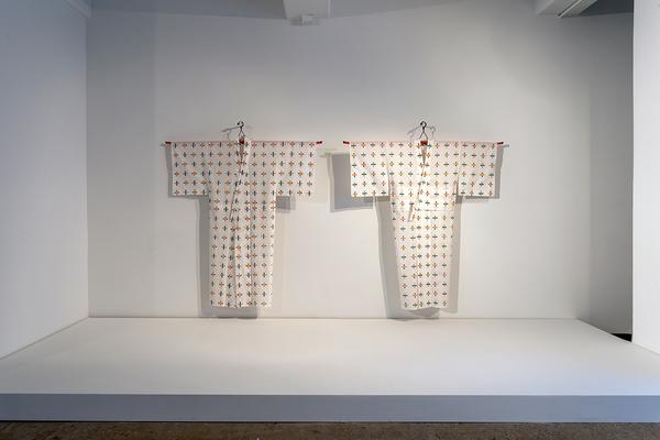 A photograph of a gallery installation featuring two white kimonos hanging side-by-side against a white wall. The kimonos are nearly identical, featuring a pattern of plus crosses with alternating marks of green and orange.