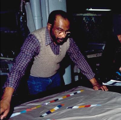 The artist Moe Brooker, a Black man wearing glasses, a plaid shirt and tan sweater vest, stands over an abstract design in progress with his arms resting on the table.  