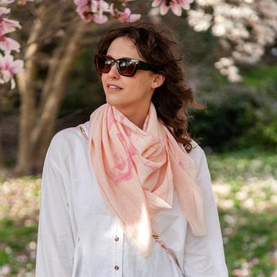 A photograph of a woman wearing a light pink scarf tied loosely around her neck, over a white blouse. She is wearing sunglasses and stands below a blossoming magnolia tree amid greenery. 