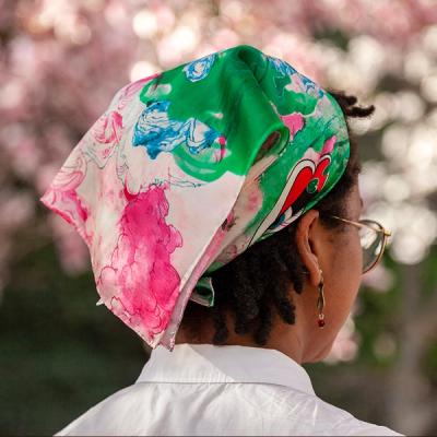 A photograph of a young Black woman turned away from the camera and wearing a small scarf around her head. The scarf features fluid ink drawings of pink, green, and blue clouds that intersect with drawings of mountains and peonies in an East Asian style. The woman is standing outdoors with the suggestion of springtime blooms in soft focus beyond her.