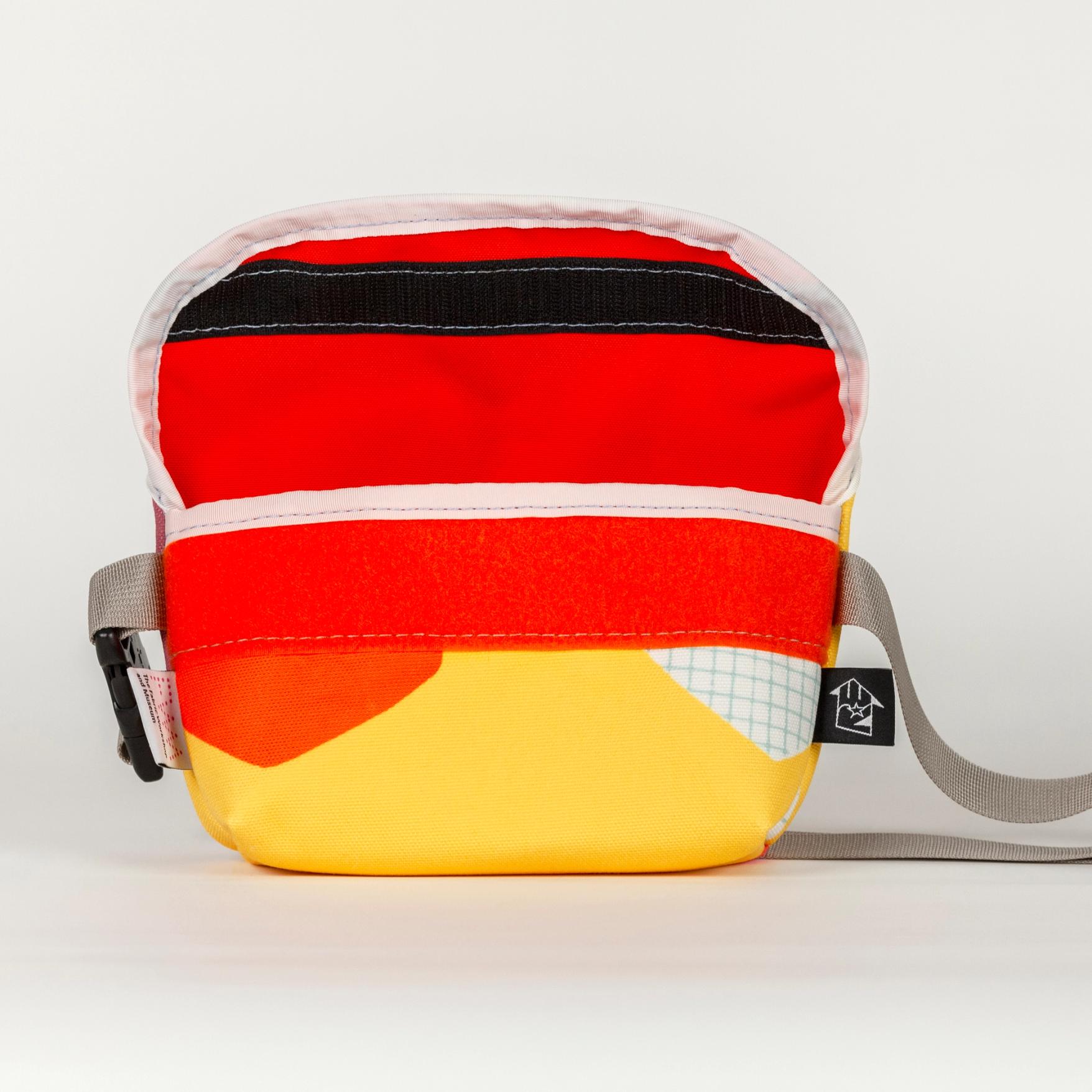 A shallow, wide, vibrant yellow bag with bold abstract shapes (bright orange and white with a light blue grid pattern). The top flap is open revealing a bright orange lining and black velcro closure with a white seam along the flap. The bag has a shiny gray strap with a plastic black buckle.