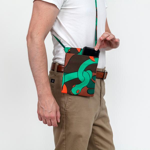 A model wearing a small rectangular cross-body bag with an exterior pocket made of fabric featuring a repeating textile pattern with stylized green snakes intertwined with abstract brown shapes and bright orange spirals on a dark brown background.