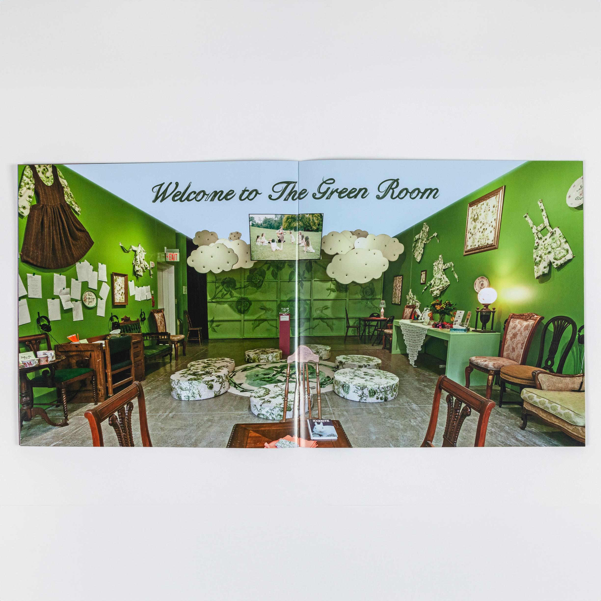 A photo of a spread of a book. There is a single photograph spanning both pages. The photograph is an installation view of "John Jarboe: The Rose Garden." The walls of the room are painted green. There is plenty of seating, mostly comprised of dining chairs made of dark wood. In the center of the room on the floor is a rug with seven large floor pillows surrounding the rug. The walls are filled with decor. The top of the photograph reads "Welcome to The Green Room"