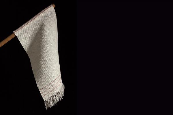 A photograph of a cream-colored dish towel hanging at a diagonal from a wooden dowel against a black background. The dishtowel has a woven texture with three thin red stripes stitched toward the frayed end.